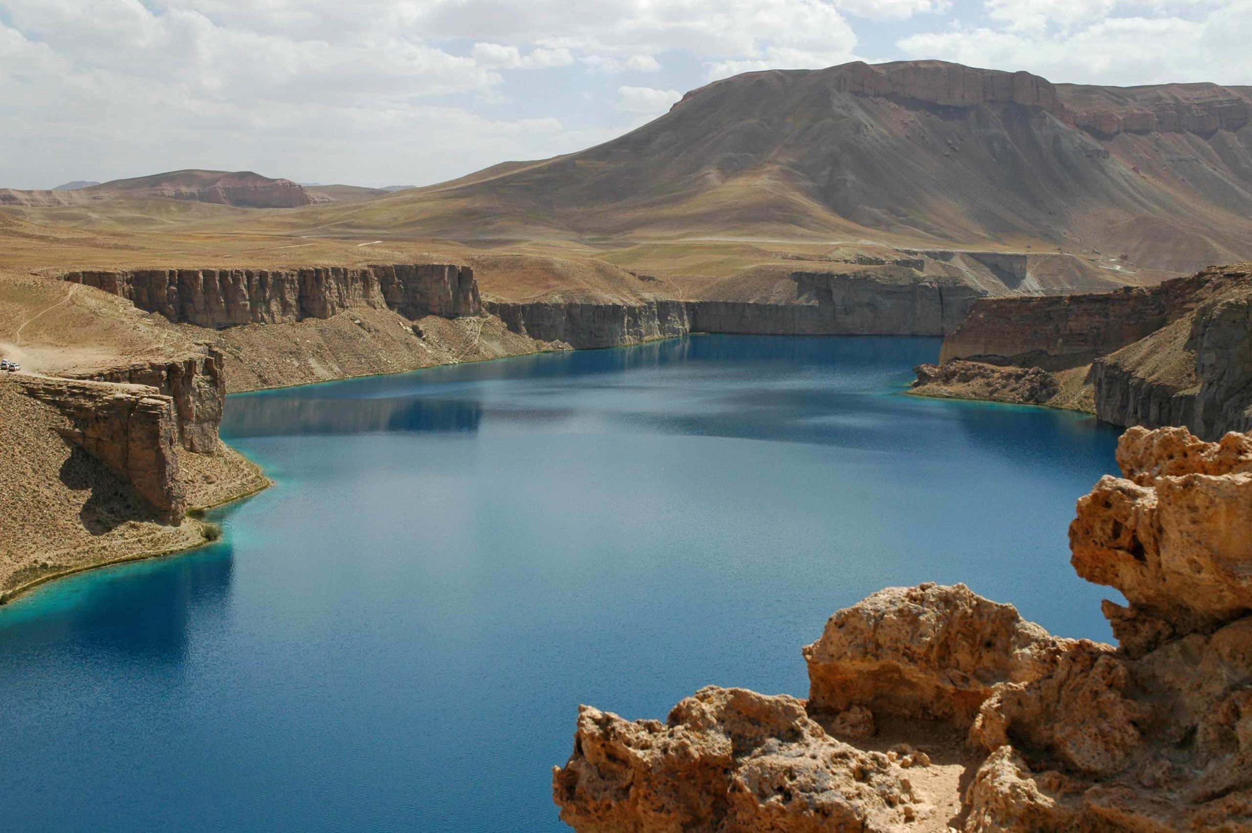 Afghanistan 桌布 壁紙 — 在部分多雲的天空下，一個令人驚嘆的藍綠色湖泊坐落在崎嶇的棕色懸崖中。