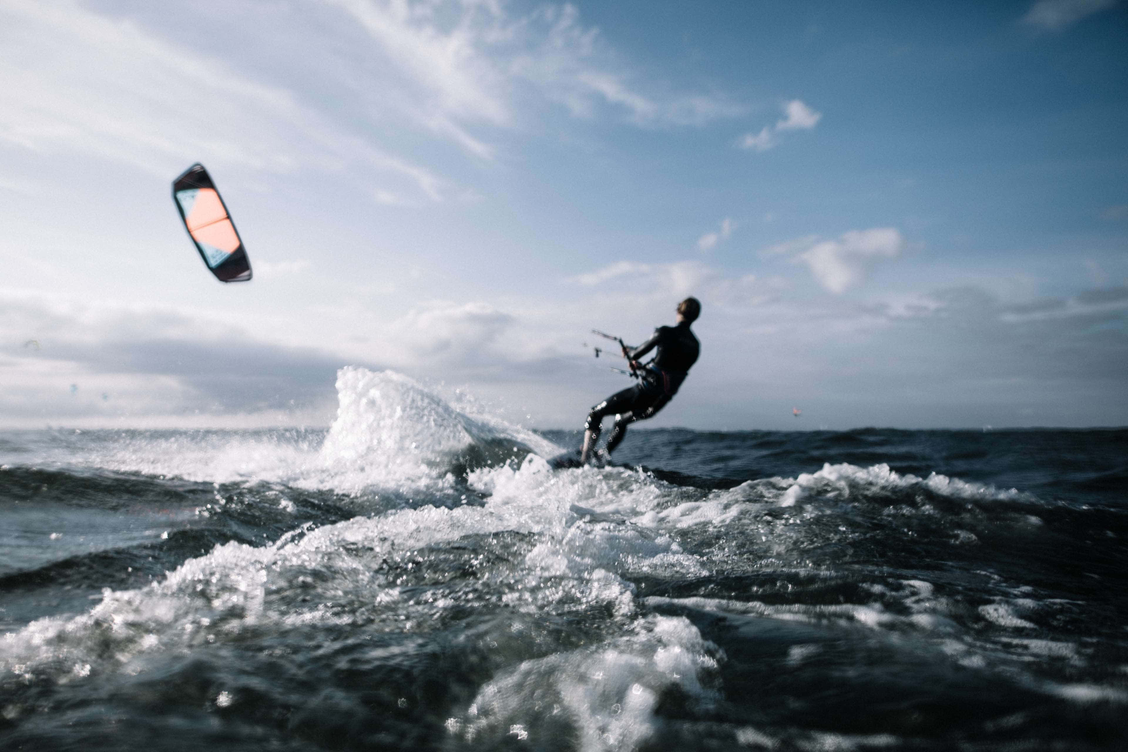 Beach Water Sports 桌布 壁紙 — 在部分多雲的天空下，一名風箏衝浪者在波濤洶湧的海面上乘風破浪，色彩繽紛的風箏在上方翱翔。