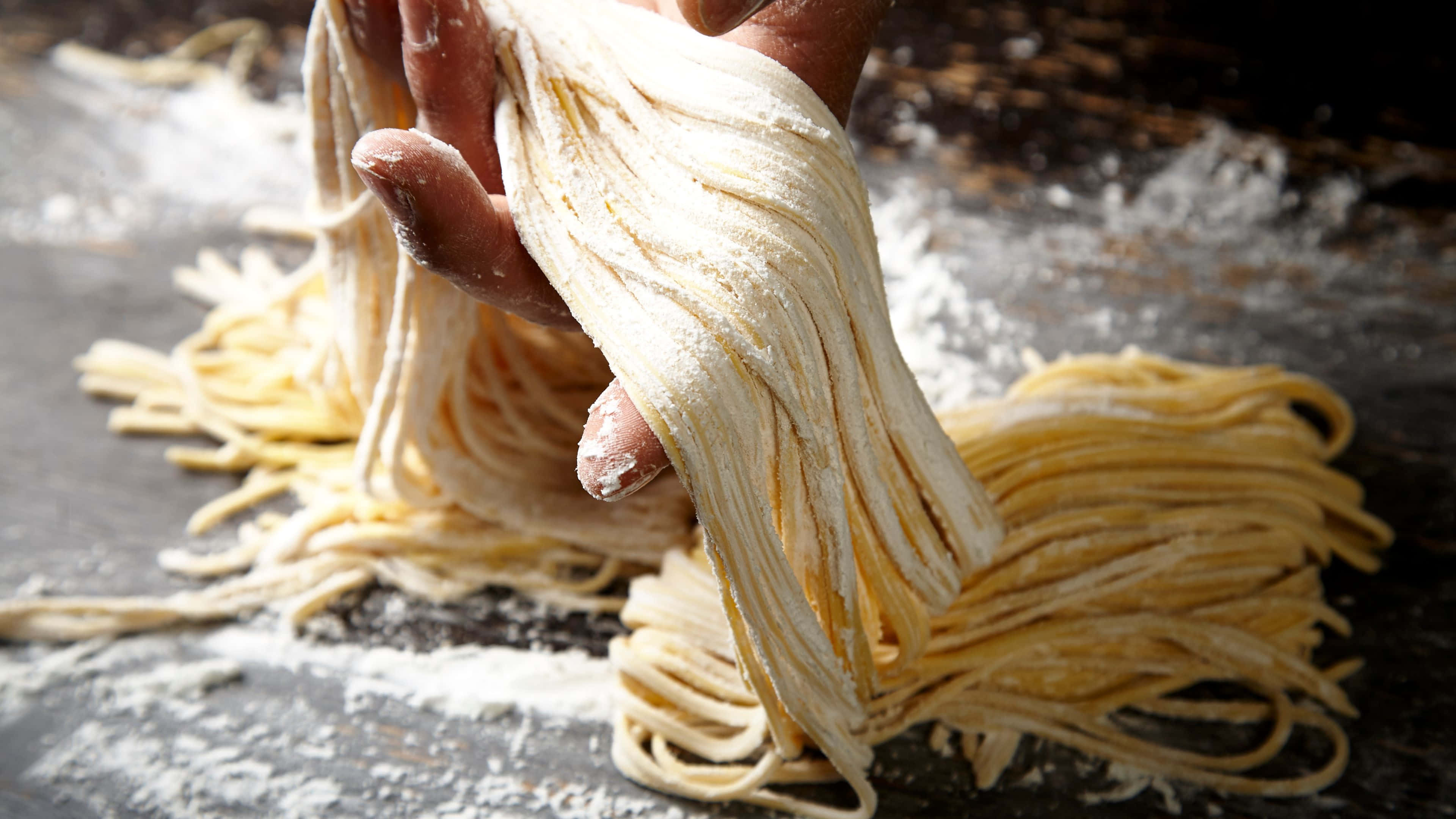 Noodles 桌布 壁紙 — 在鄉村風格的廚房環境中，一隻手從撒了麵粉的木質表面上拉出新鮮的手工意粉條。