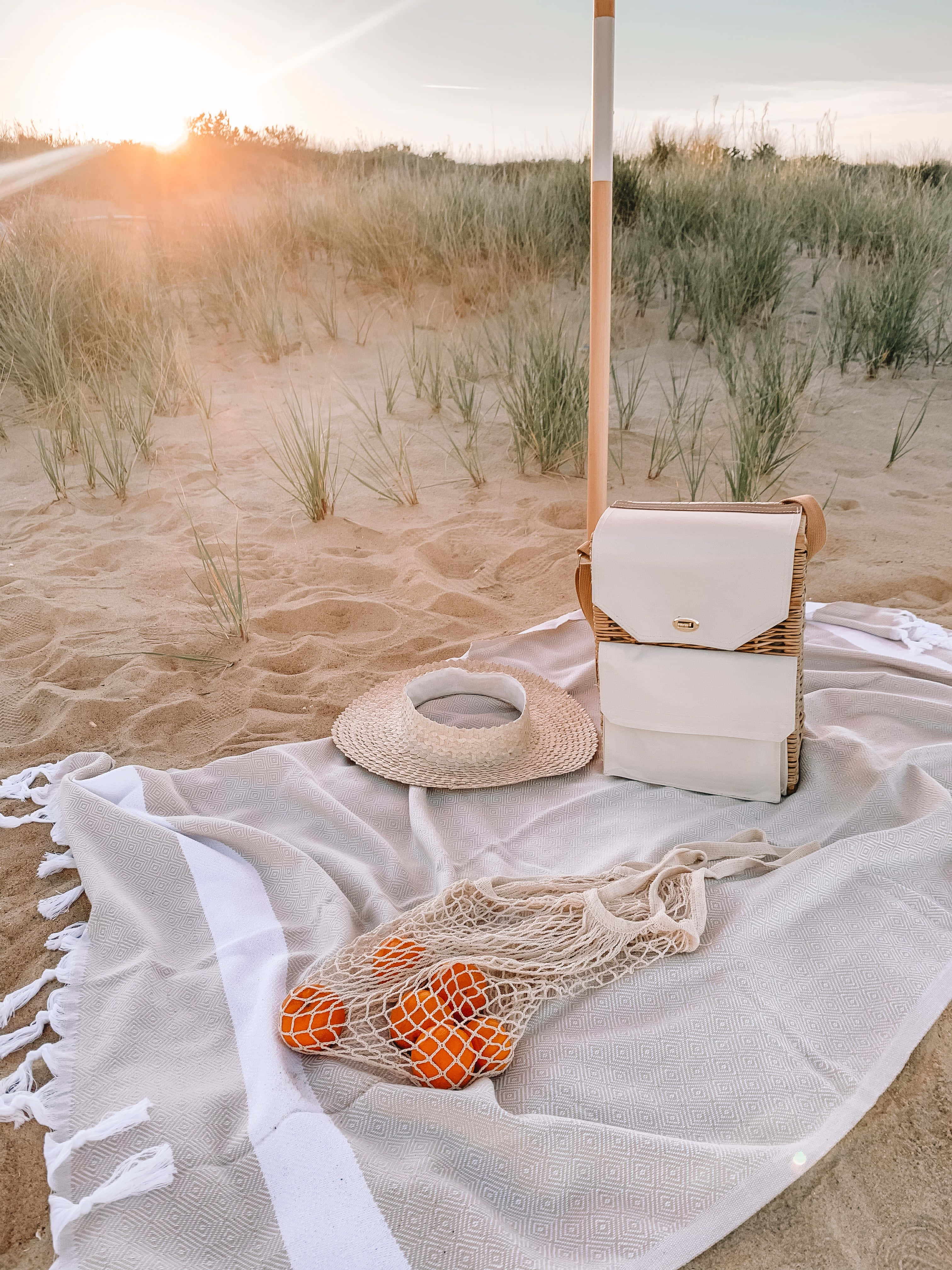 Beach Picnic 桌布 壁紙 — 在金色日落時分，一個寧靜的海灘野餐佈置，有草帽和橙子放在圖案毯子上。