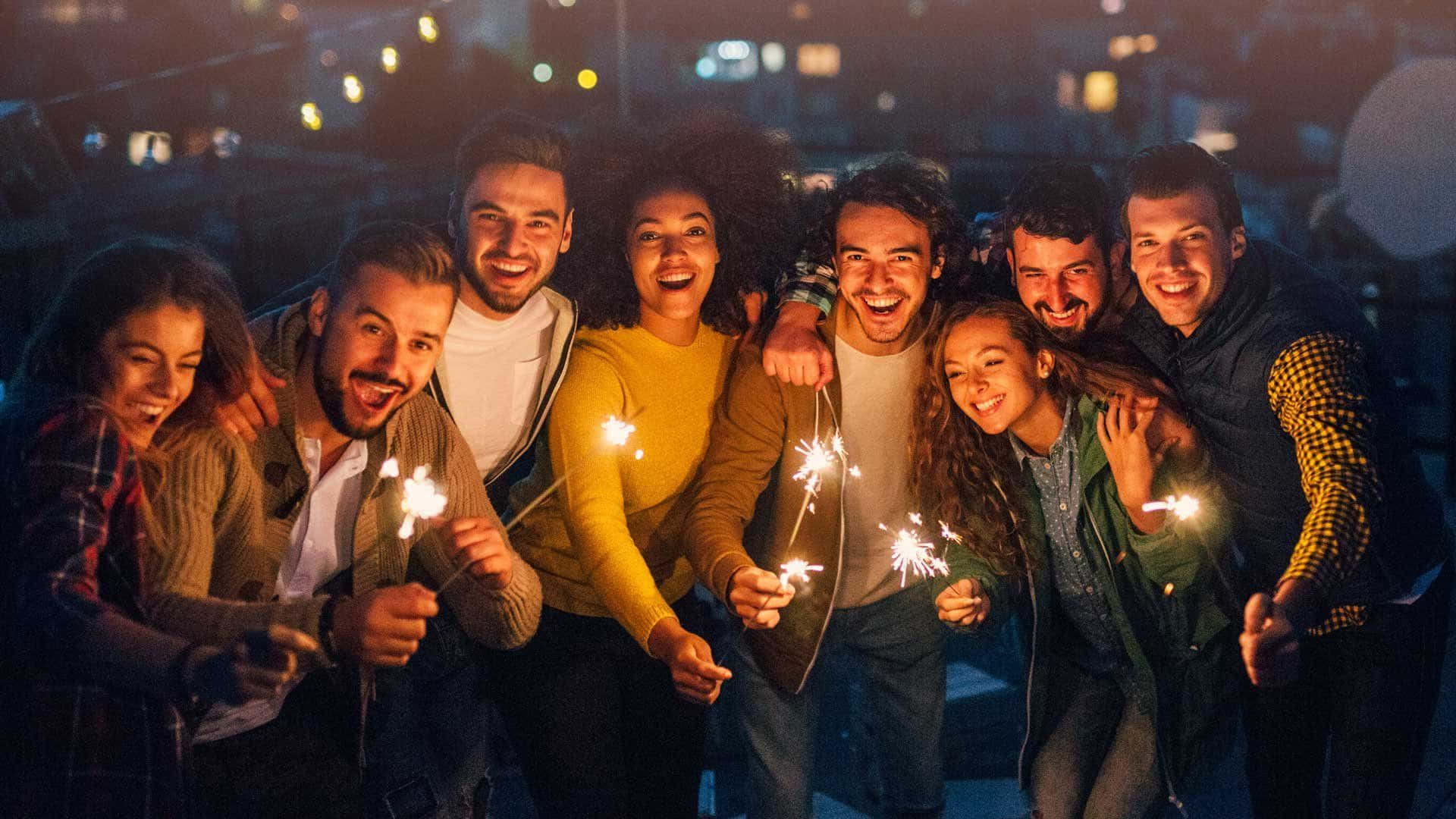 在閃爍燈光下熱鬧的派對夜晚 壁紙 — 一群快樂的朋友在晚上於屋頂上大笑並手持煙花棒，背後的城市燈光閃爍。