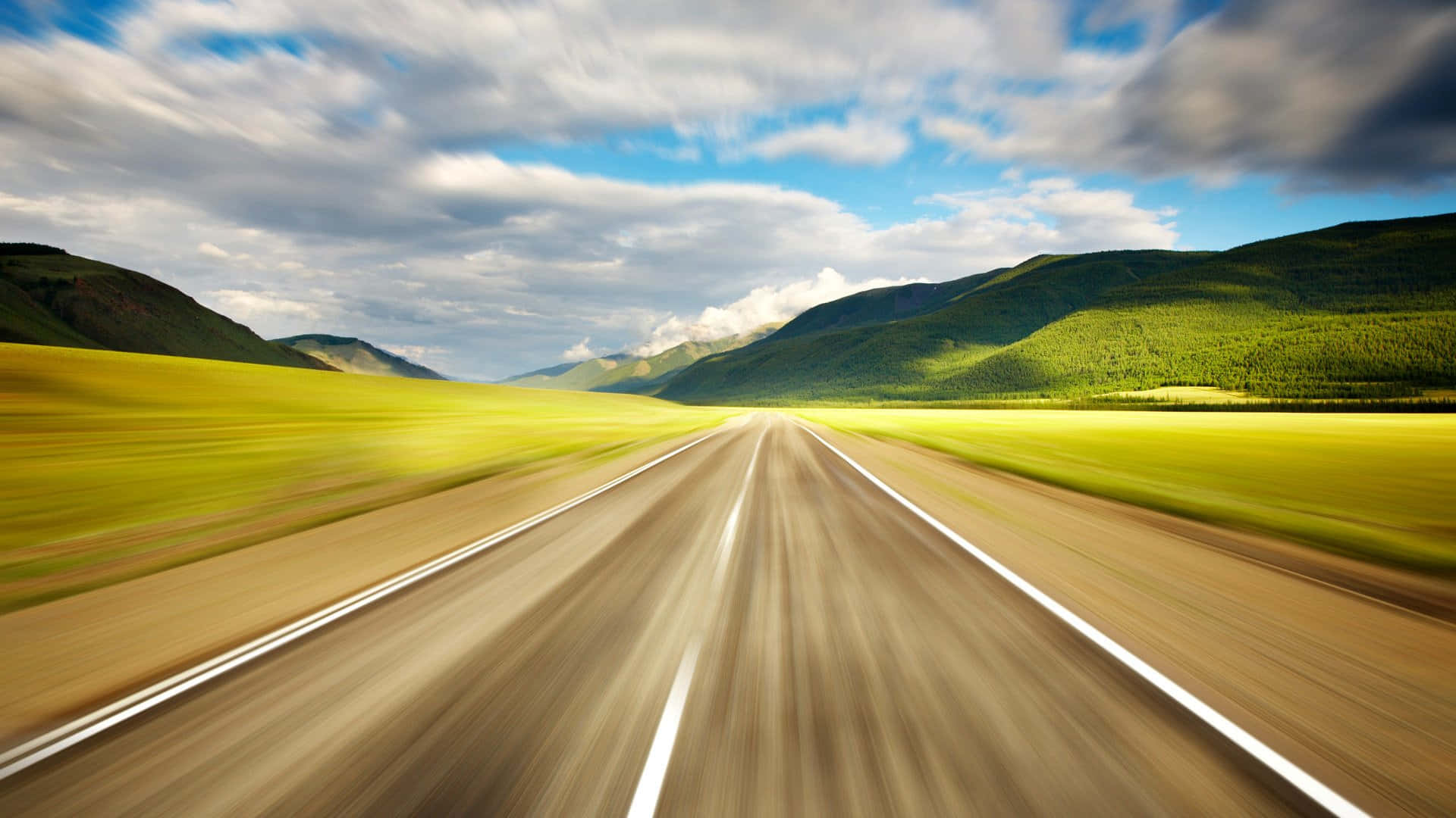 在開闊道路上啟程 壁紙 — 在動態多雲的天空下，一條筆直的道路兩旁是充滿活力的黃色田野和綠色山脈，高速行駛的模糊視野。