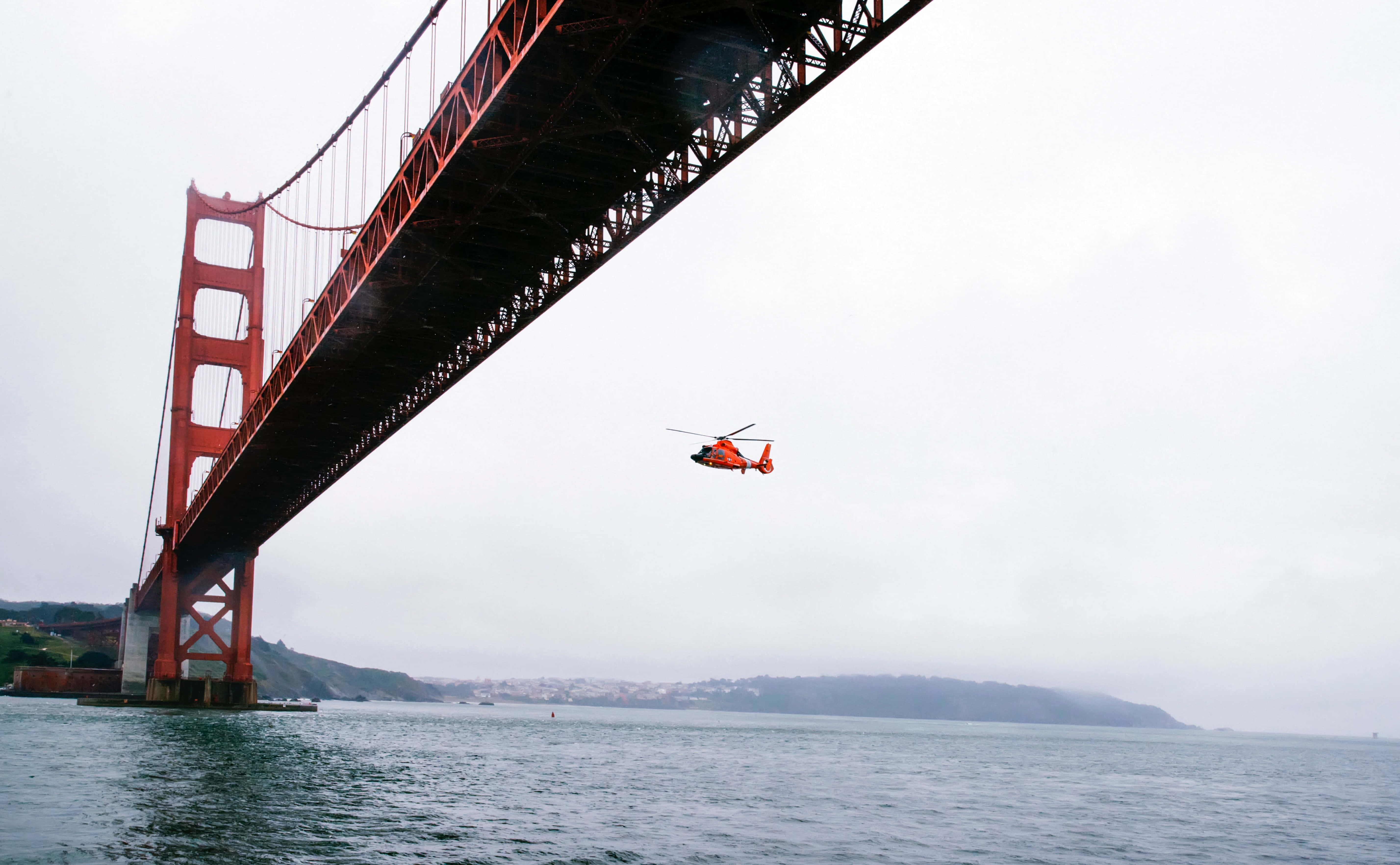 直升機 桌布 壁紙 — 在陰天、背景有霧氣籠罩的山丘下，一架亮橙色的直升機飛越標誌性的金門大橋。