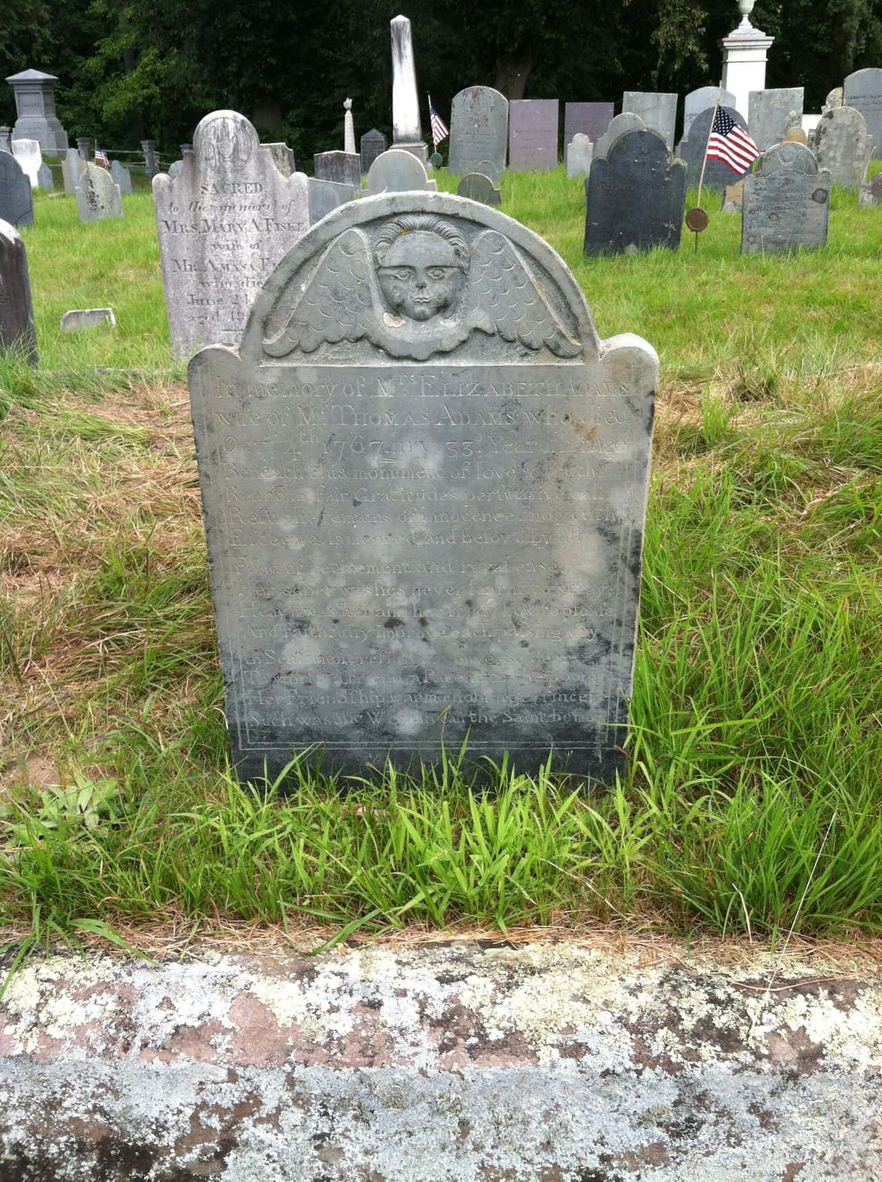 Gravestone 桌布 壁紙 — 在陰天之下，一個長滿草的墓園中，有一座風化的灰色墓碑，上面雕刻著天使般的面孔。