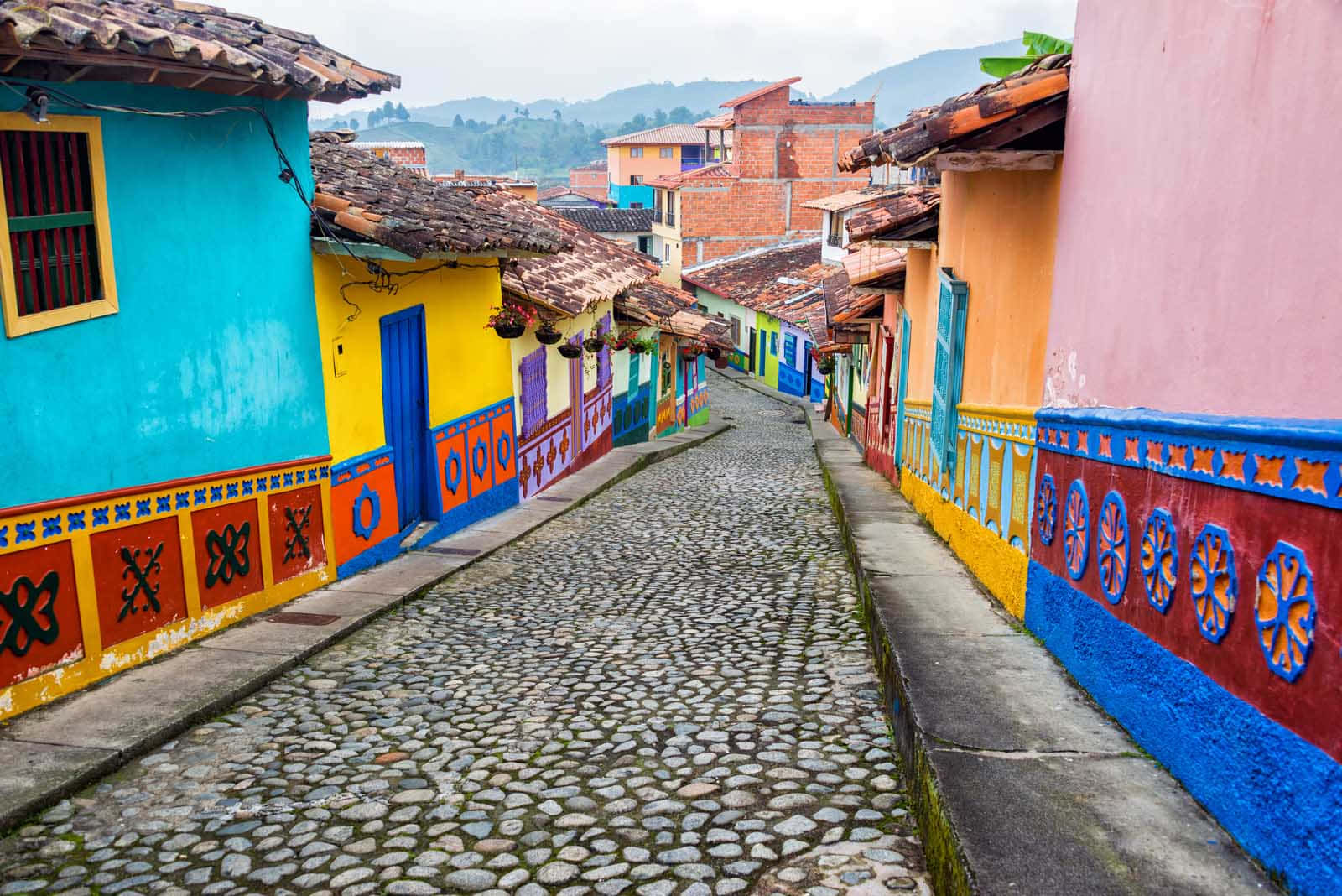 Colombia 桌布 壁紙 — 在陰天之下，一條充滿活力的鵝卵石街道兩旁是色彩繽紛的殖民風格房屋，帶有圖案壁畫。
