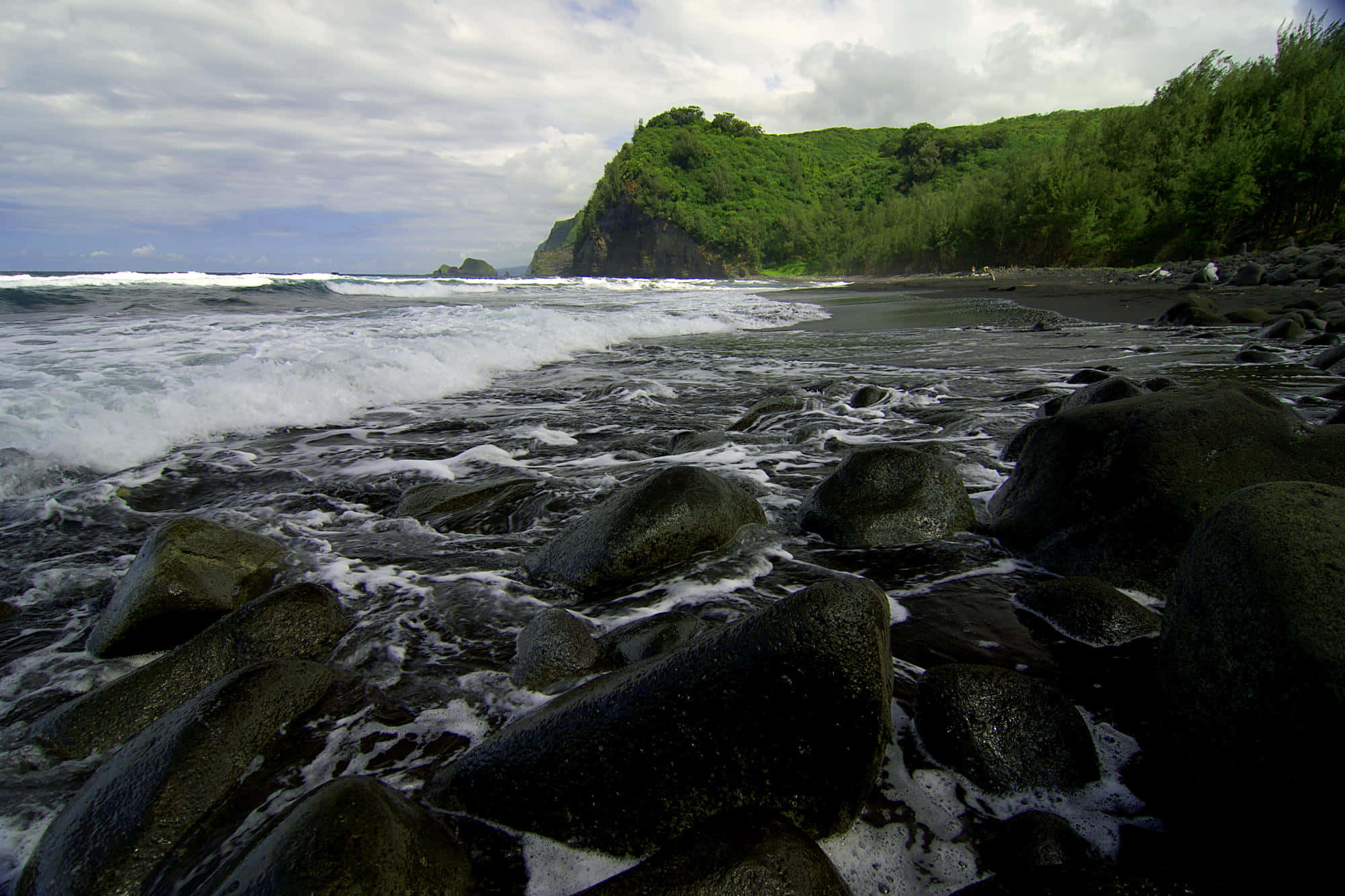 Black Sand Beach 桌布 壁紙 — 在陰天之下，海浪拍打著黑色沙灘上的深色火山岩，周圍是茂密的綠色懸崖。