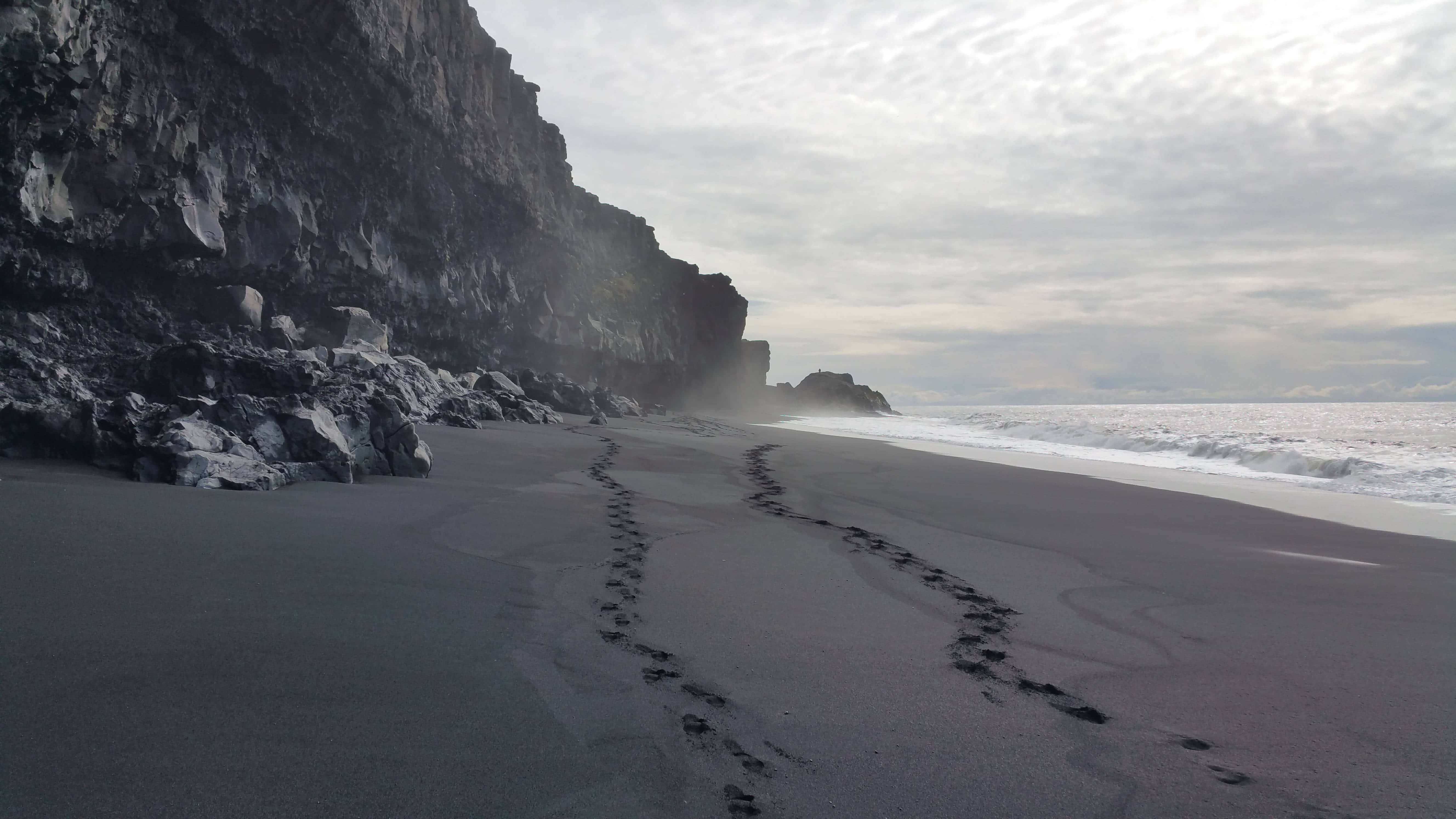 Black Sand Beach 桌布 壁紙 — 在陰天之下，腳印沿著霧氣瀰漫的黑沙灘延伸，旁邊是高聳的火山懸崖。