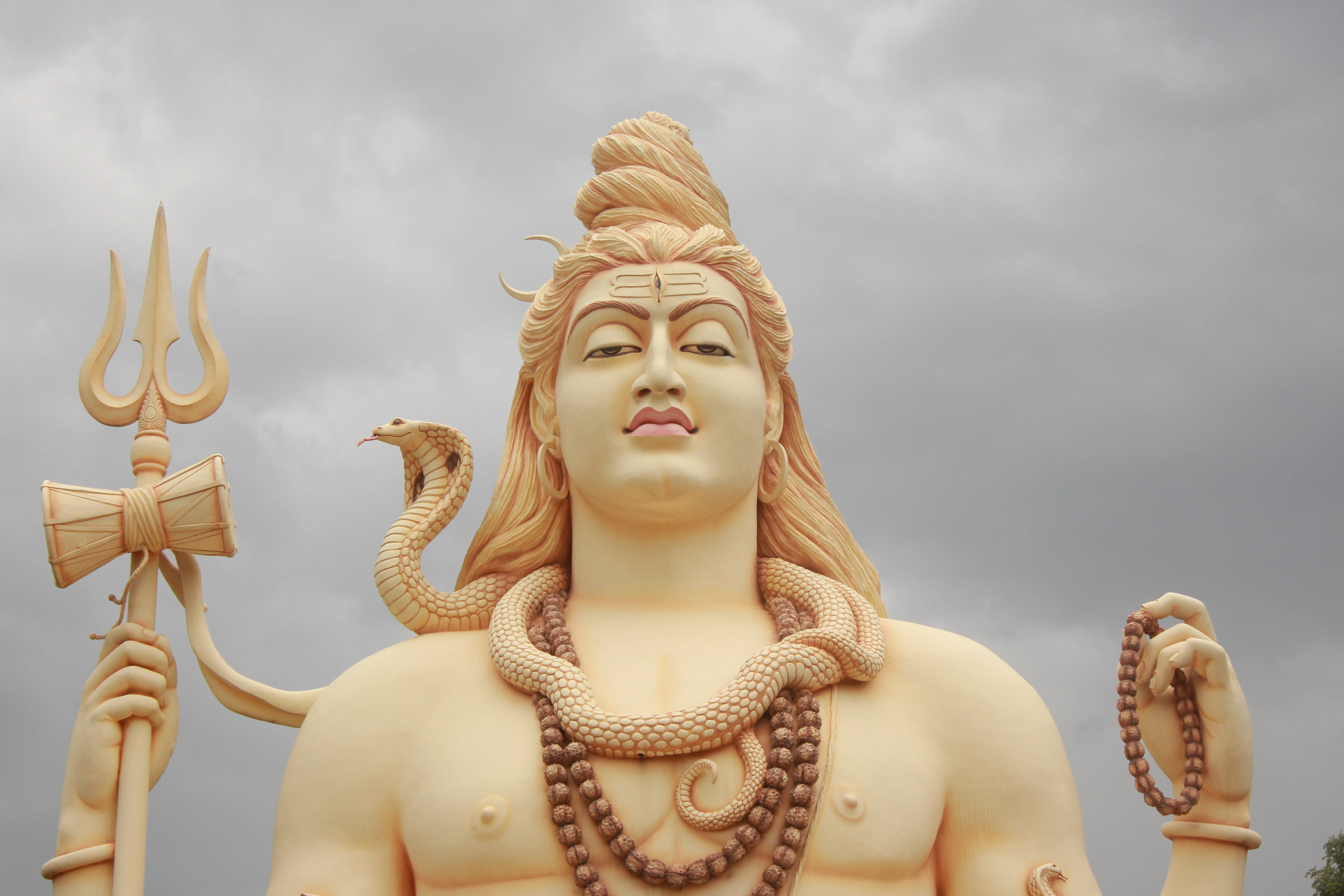 Lord Shiva 桌布 壁紙 — 在陰天天空下，一座高聳的濕婆神雕像，手持三叉戟、蛇和念珠。