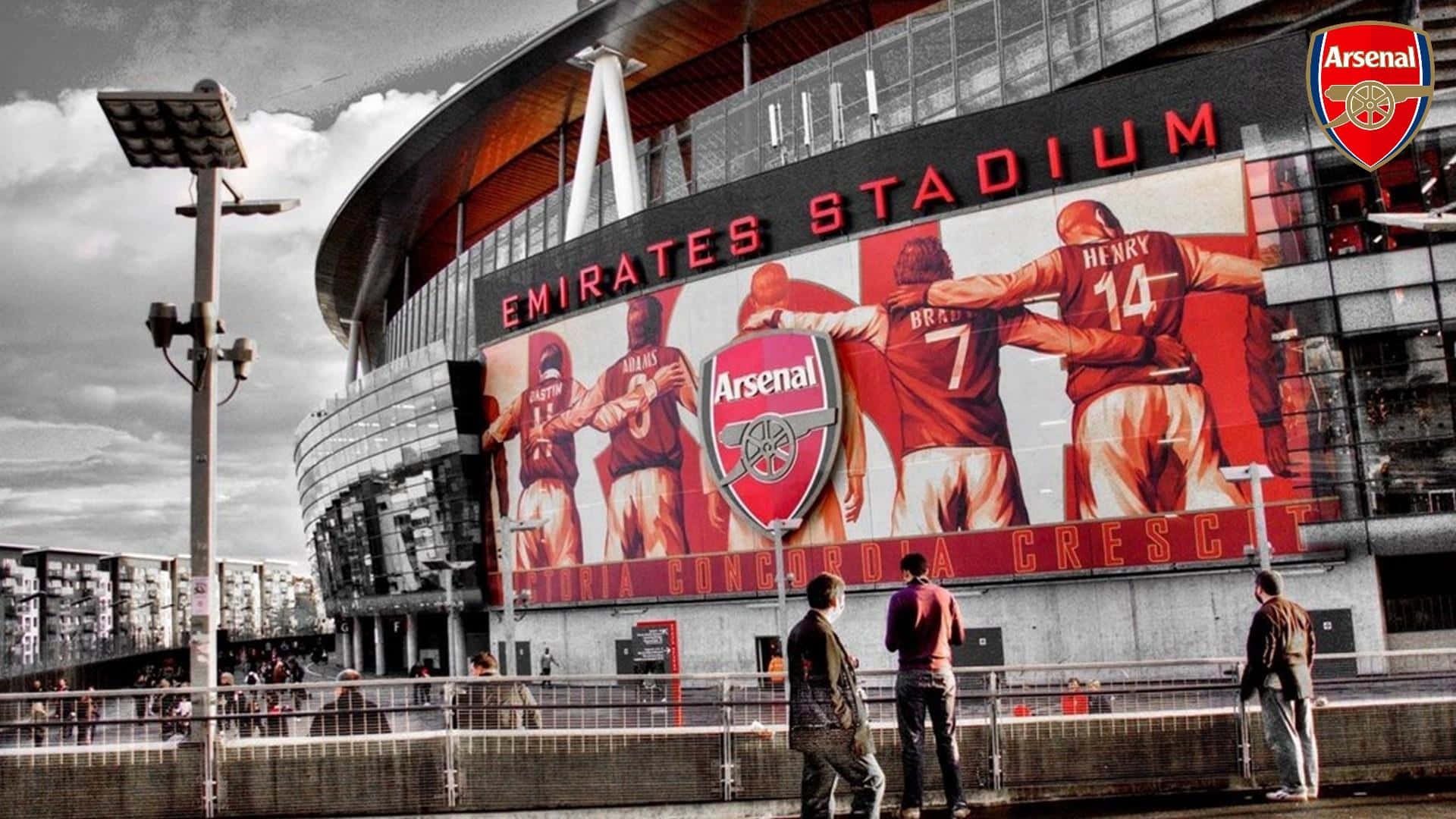 Arsenal 桌布 壁紙 — 在陰天天空下，酋長球場戲劇性的外觀，帶有醒目的紅色球員壁畫和下方行走的球迷。
