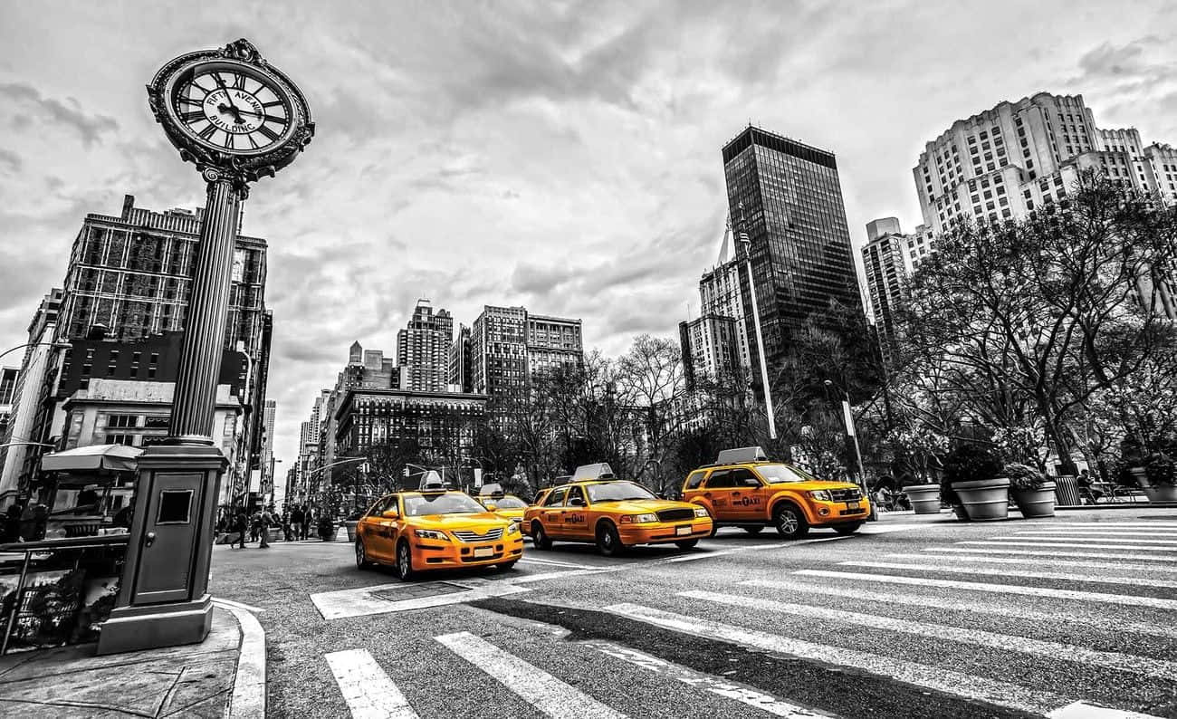 Yellow Cab 桌布 壁紙 — 在陰天天空下，鮮黃色的的士與黑白城市景觀形成對比，城市景觀中有一座高聳的時鐘和摩天大樓。