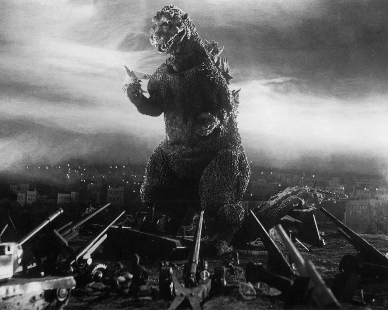 Godzilla 1954 桌布 壁紙 — 在陰雲密布的天空下，一隻高聳的哥斯拉站立於黑白城市景觀中被摧毀的坦克和瓦礫之中。
