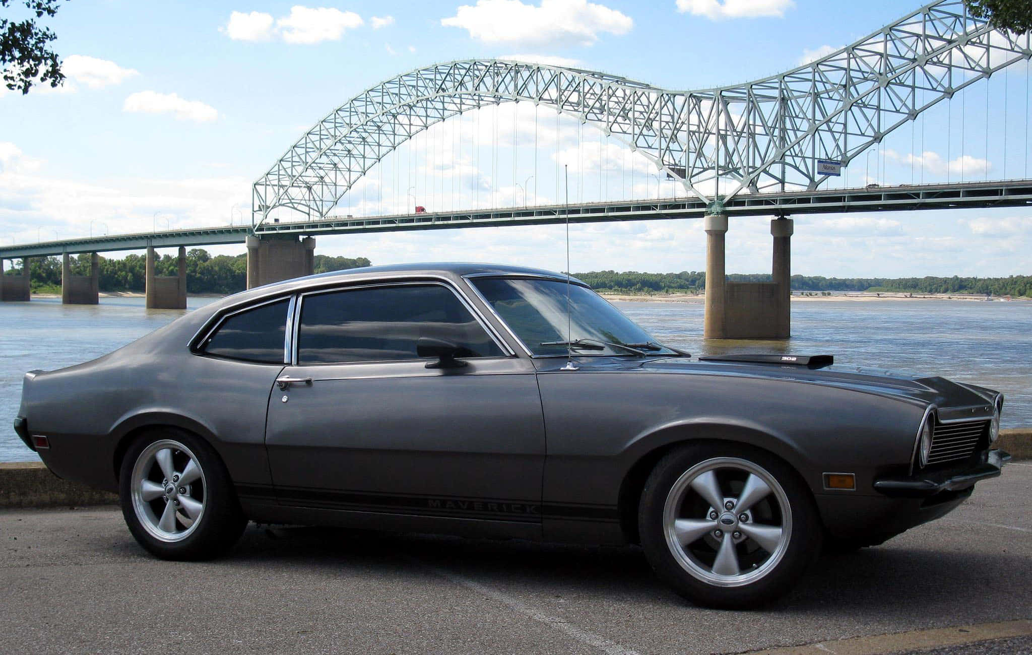 Ford Maverick 桌布 壁紙 — 在陽光明媚的日子裡，一輛時尚的灰色經典肌肉車停泊在鋼拱橋下的河邊。