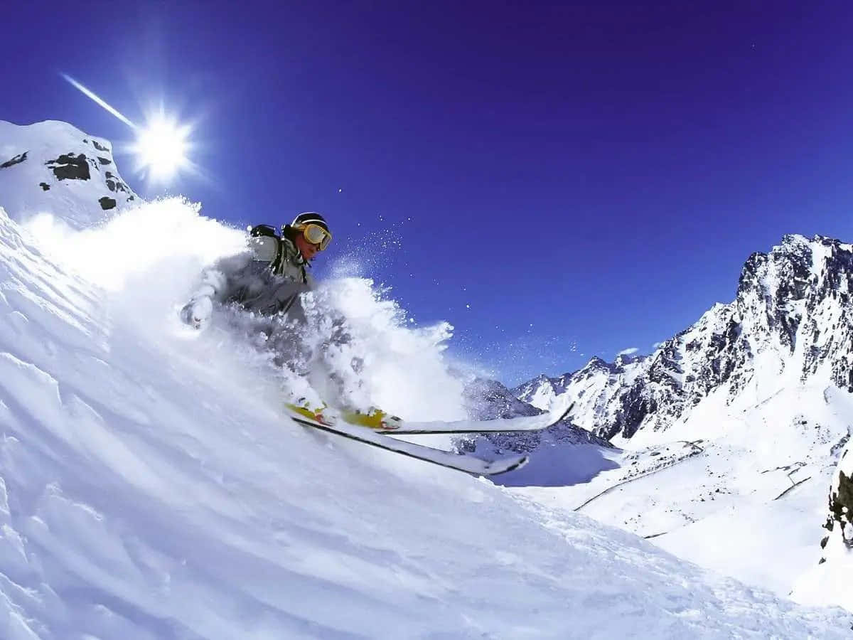 Skiing 桌布 壁紙 — 在陽光普照的雪坡上，一名滑雪者在湛藍天空下劃過新鮮粉雪，周圍是鋸齒狀的山峰。