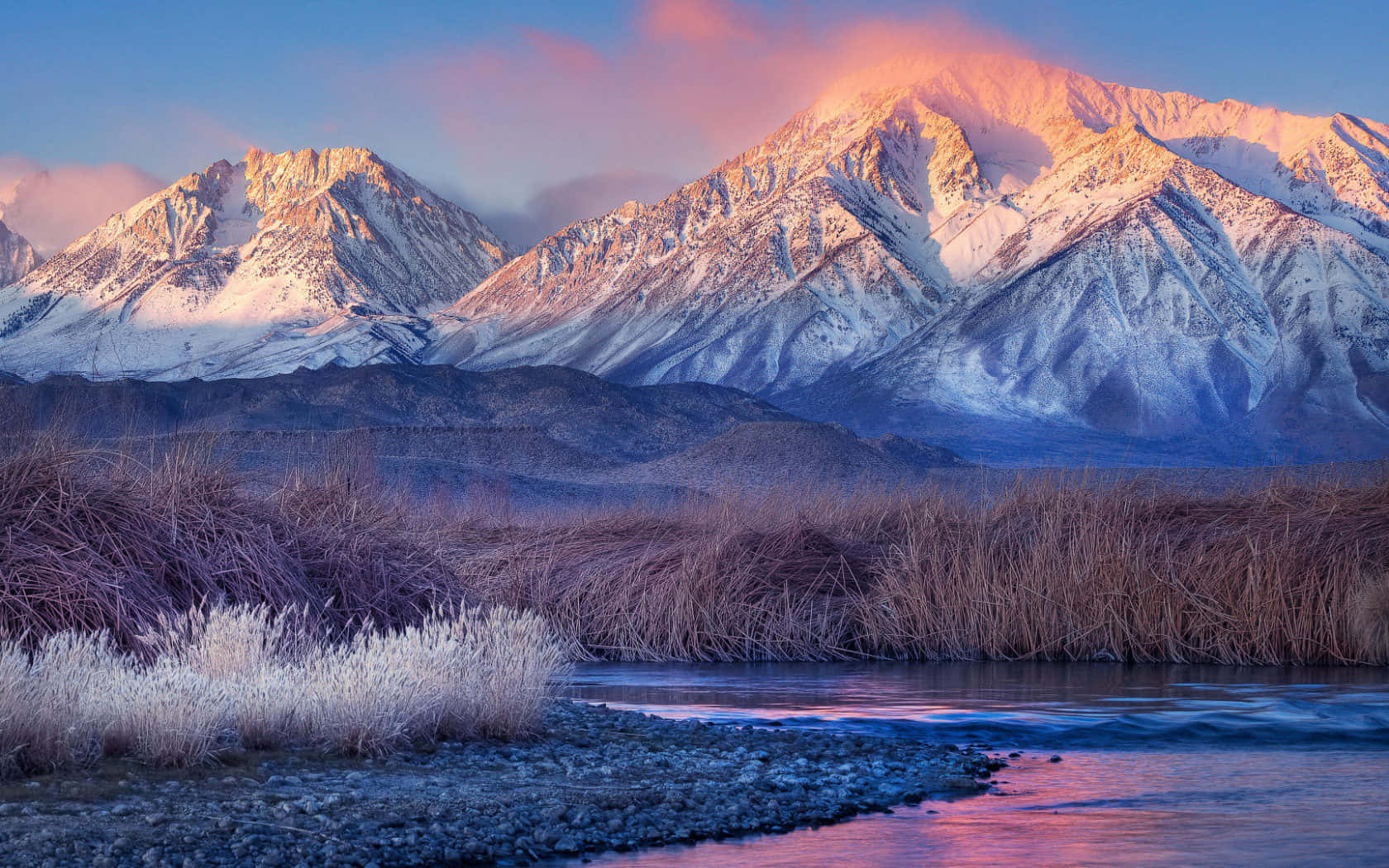 在雄偉雪山之巔感受自然奇觀 壁紙 — 積雪的山峰在日出時閃耀著粉紅色和金色，河流在結霜的蘆葦下倒映著天空。