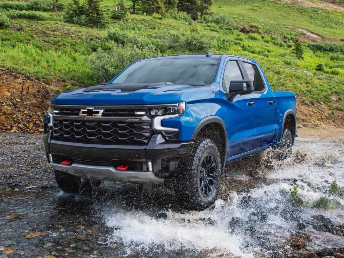 在雪佛蘭中感受道路的澎湃力量！ 壁紙 — 一輛充滿活力的藍色 Chevrolet Silverado 卡車在茂密的綠色山地地形中濺起水花穿過岩石溪流。