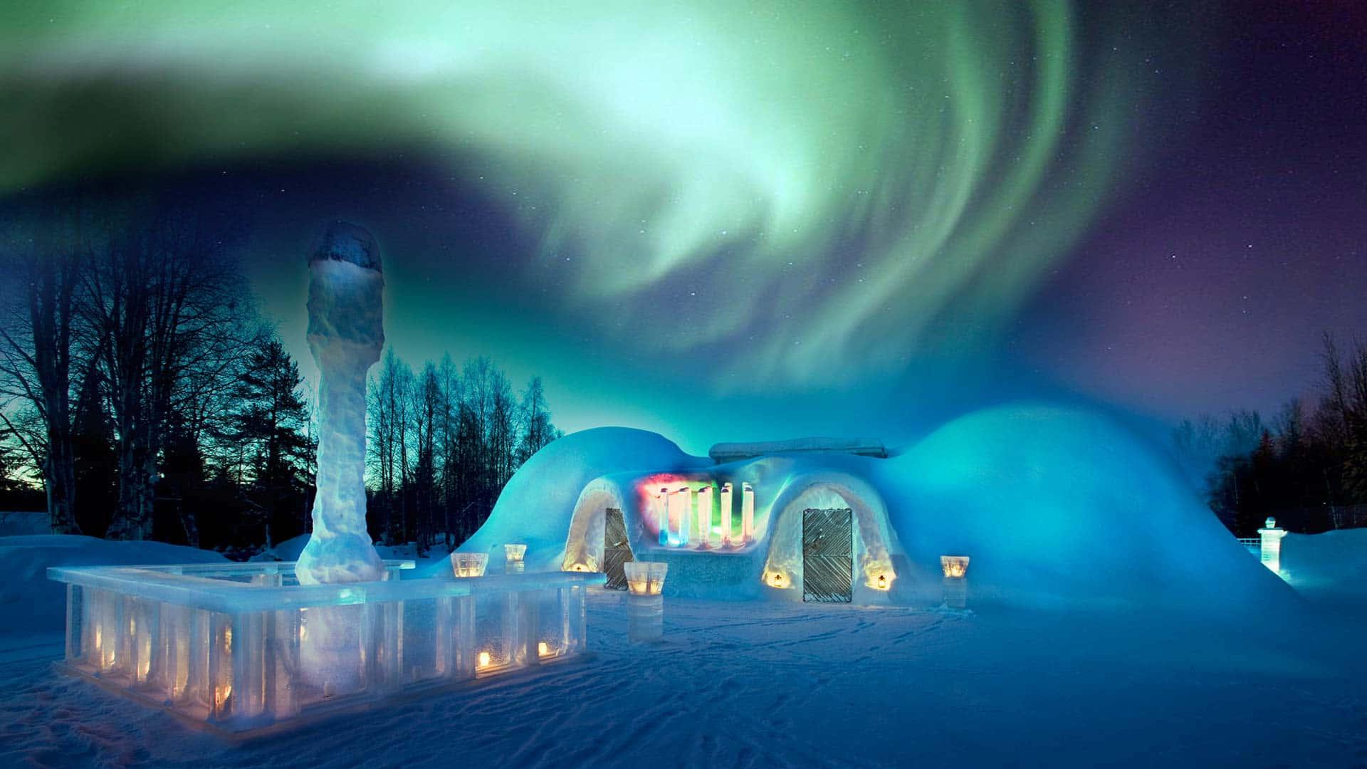 Ice Hotel 桌布 壁紙 — 在雪夜森林中，一座亮燈的冰酒店在旋轉的綠色北極光下閃耀。
