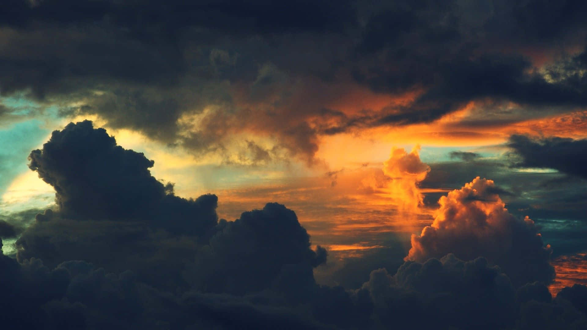 在雲朵間尋得寧靜，感受美學雲景 壁紙 — 戲劇性的日落將暴風雲染成火橙色和深藍綠色，光線穿過動盪的天空。