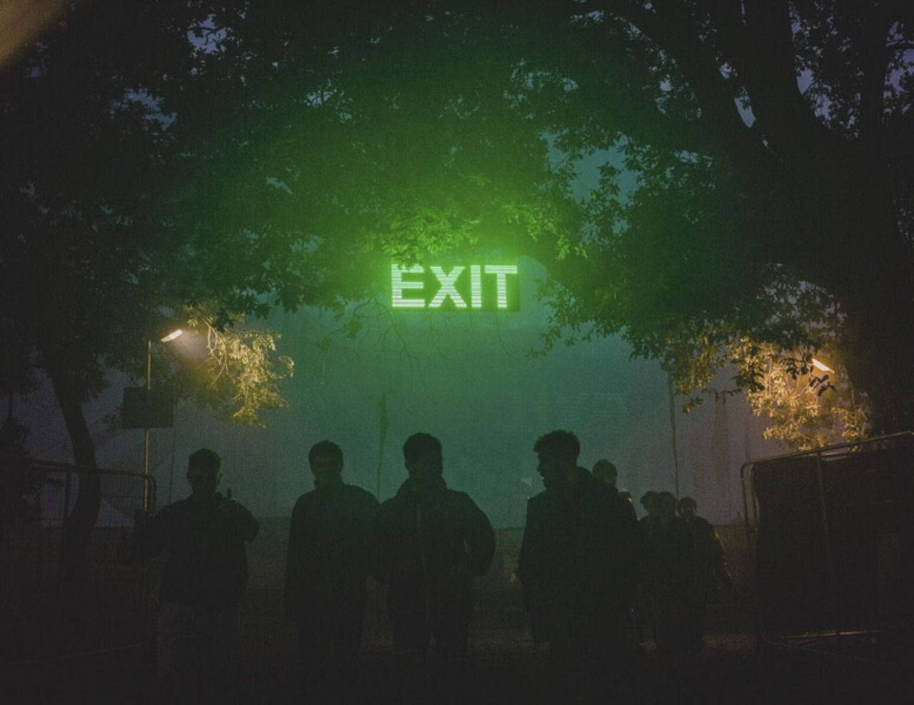 Silhouette 桌布 壁紙 — 在霧氣瀰漫的夜晚場景中，樹木環繞的發光綠色出口標誌下方站著剪影人物。