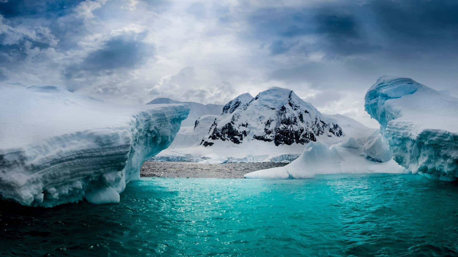 Antarctica 桌布 壁紙 — 在風暴天空下，高聳的冰山框住碧綠海洋的戲劇性北極場景。