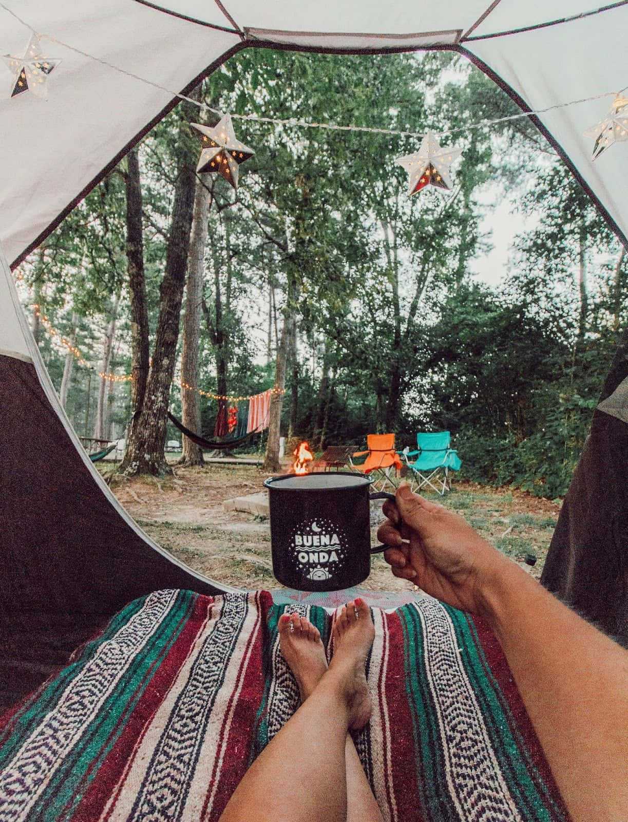Winter Camping 桌布 壁紙 — 在黃昏的帳篷內，一個人拿著杯子，赤腳放在彩色毯子上，外面可見森林和營火。