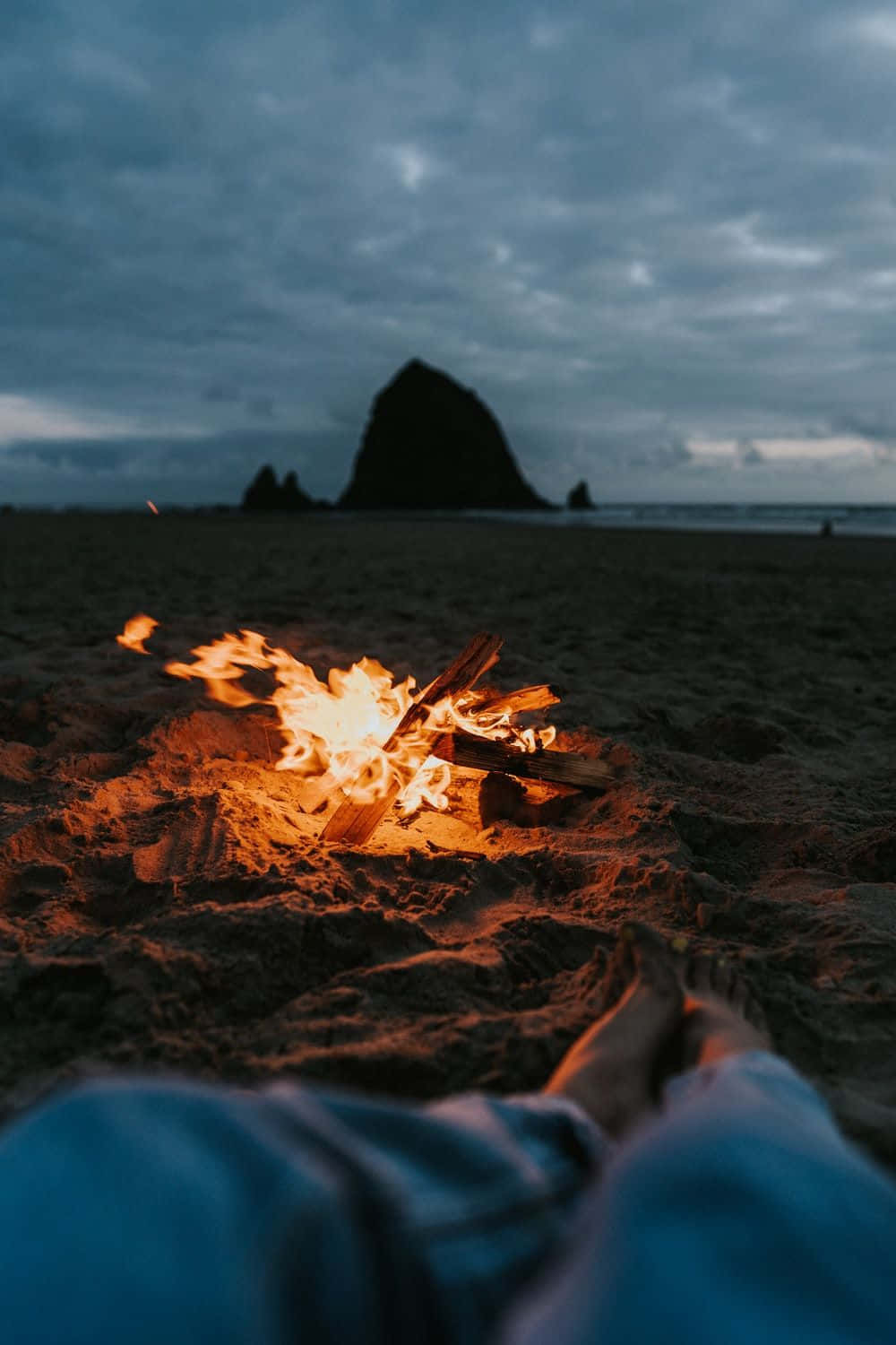 Beach Bonfire 桌布 壁紙 — 在黃昏的沙灘上，溫暖的營火明亮地燃燒，附近有人的赤腳，遠處有海蝕柱的剪影。
