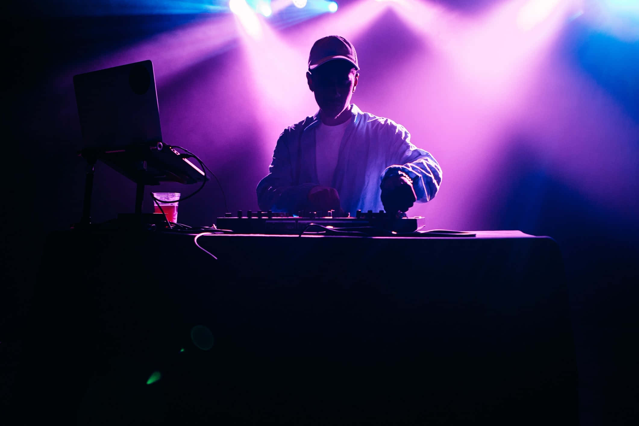 DJ 桌布 壁紙 — 在黑暗的俱樂部環境中，一個剪影的DJ在充滿活力的紫色和藍色舞台燈光下混音曲目。