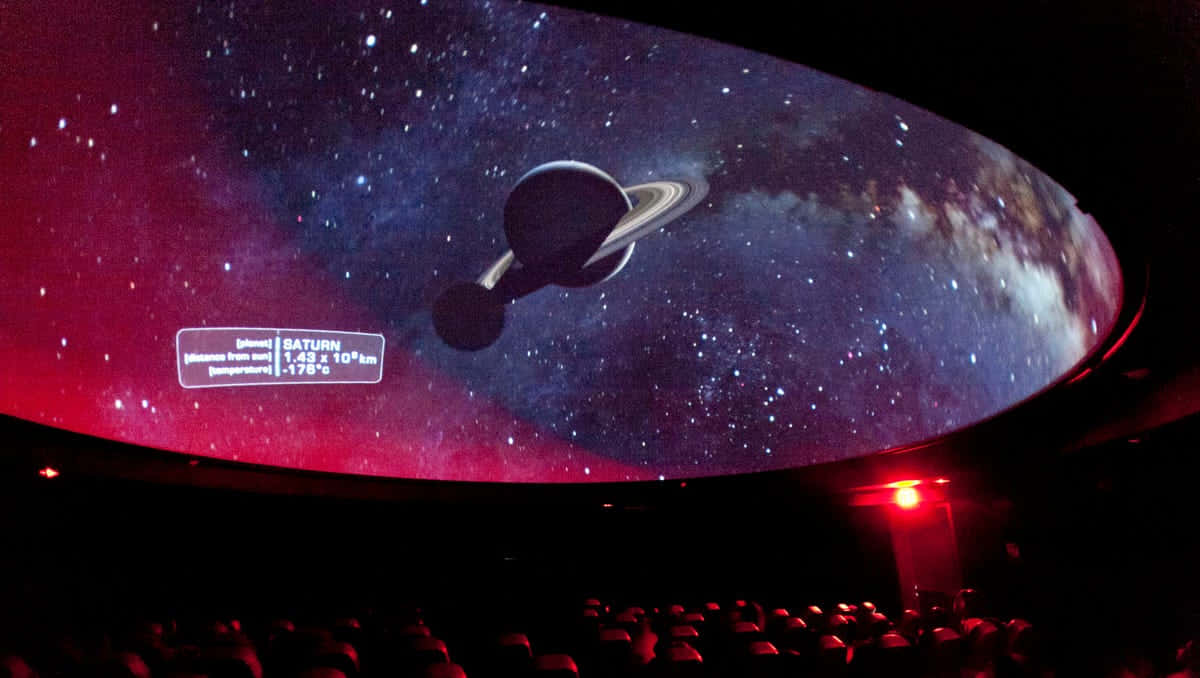 Planetarium 桌布 壁紙 — 在黑暗的劇院中，彎曲的圓頂屏幕顯示帶有光環的土星，背景是繁星點點的宇宙，並有數據疊加。