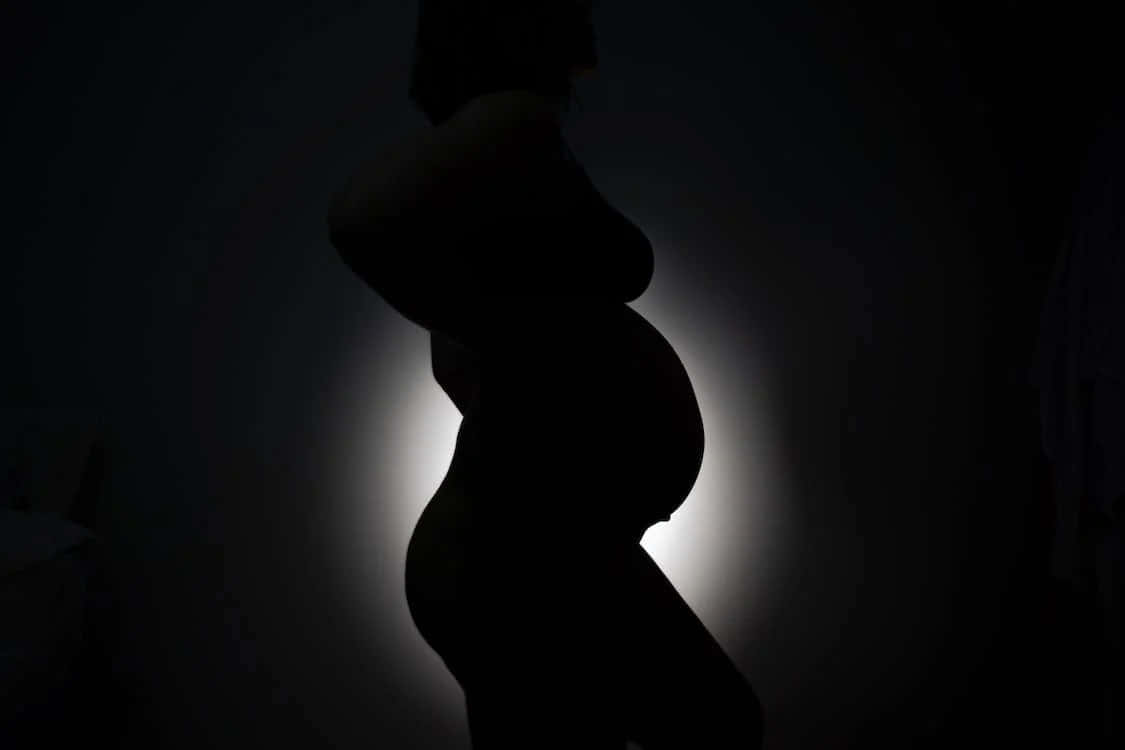 pregnancy 桌布 壁紙 — 在黑暗的房間中，孕婦的剪影映襯著柔和發光的背光。