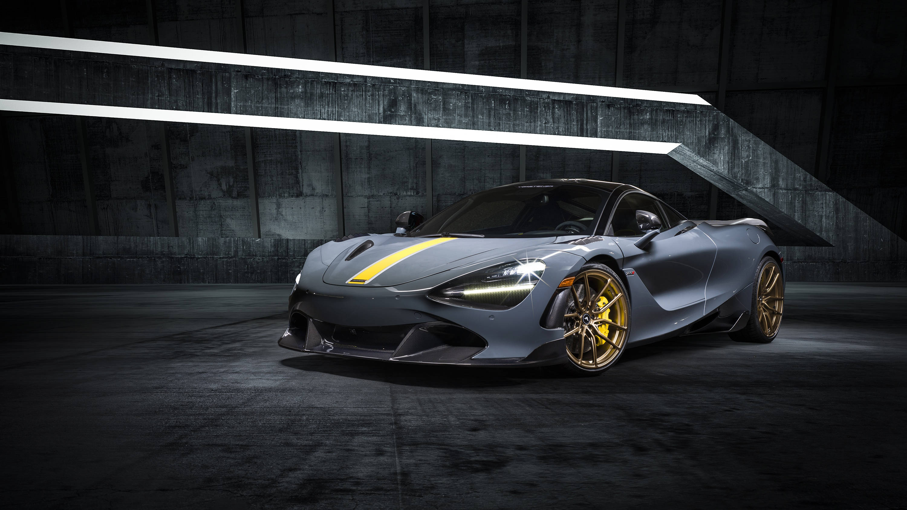 地下停車場的麥拉倫720S 壁紙 — 一輛光滑的啞光灰色McLaren 720S，帶有金色飾邊和黃色條紋，在黑暗的混凝土車庫中，在刺眼的白光下閃耀。