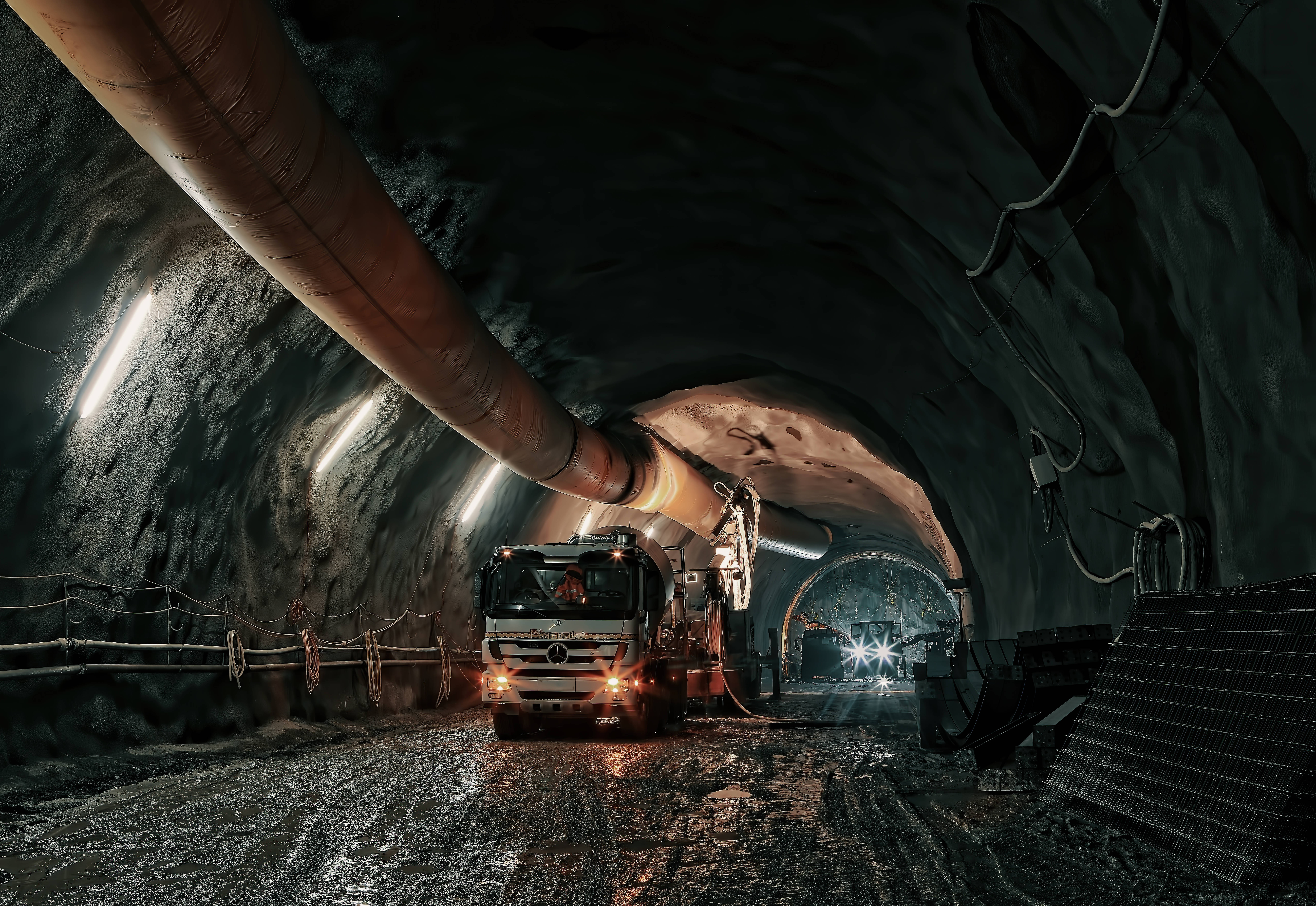 地下採礦 壁紙 — 一架重型礦用卡車在光線昏暗的地下隧道中行駛，隧道內有裸露的岩壁和頭頂的工業管道。