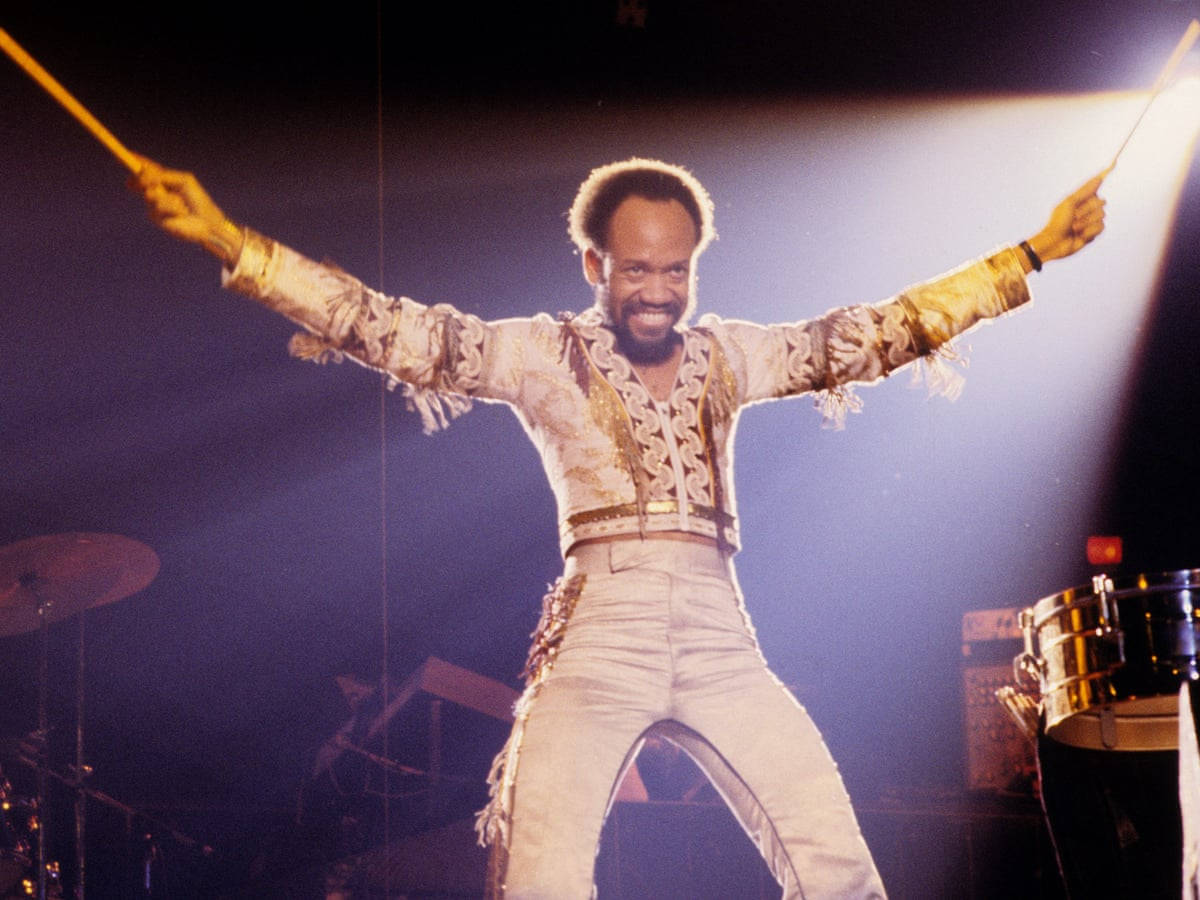 地球、風與火樂隊莫里斯·懷特手持鼓棒 壁紙 — 一位身穿閃亮金色服裝的快樂鼓手在明亮的聚光燈下張開雙臂跳上舞台。