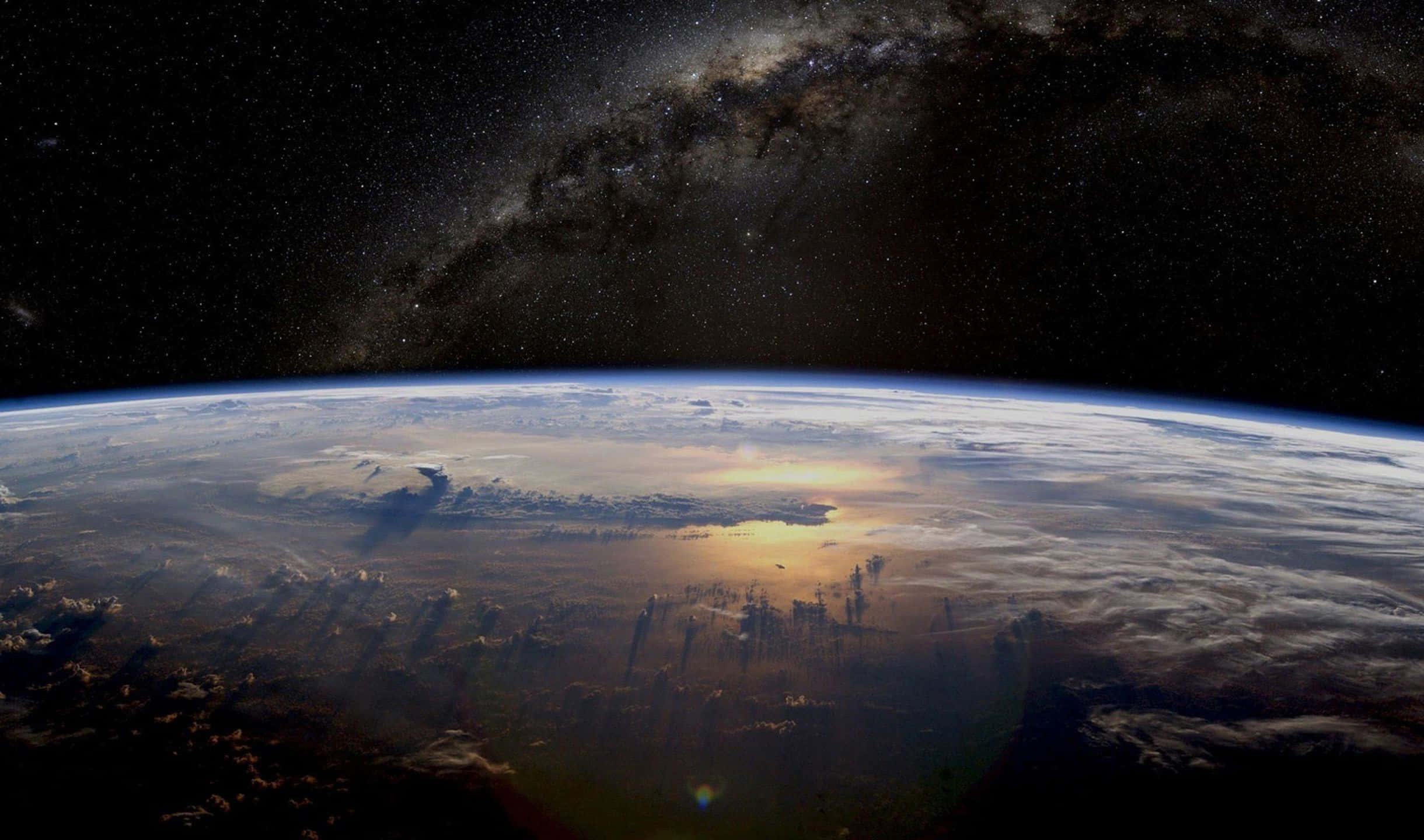 2440x1440 桌布 壁紙 — 地球在星空下彎曲，銀河在頭頂拱起，陽光在雲層上閃爍。