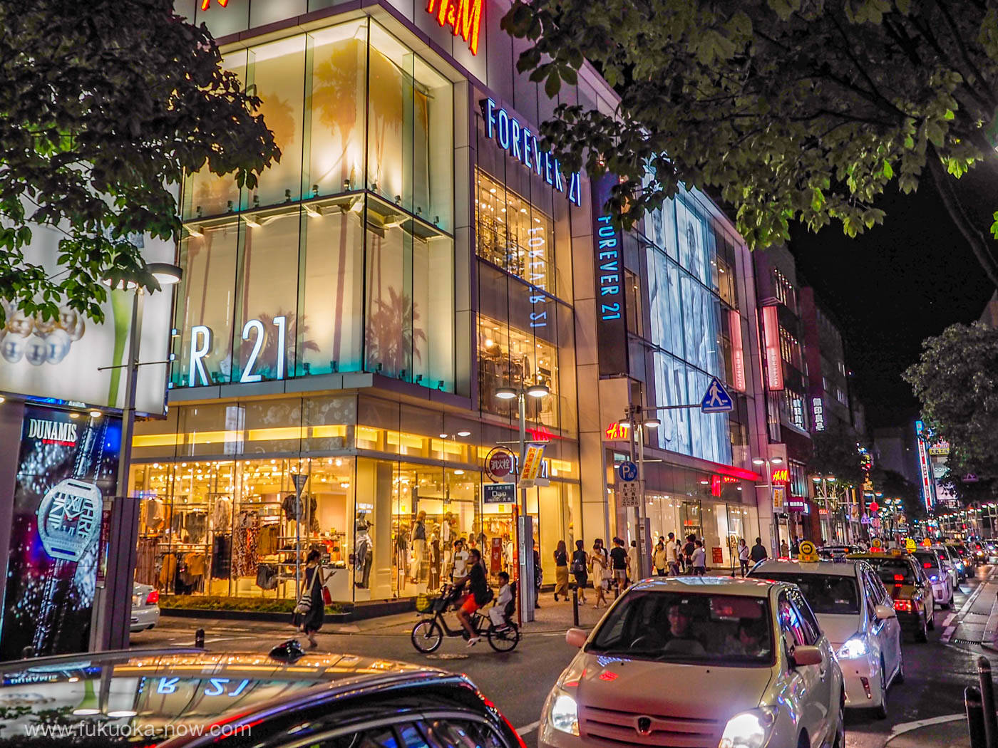 地鐵旁的 Forever 21 壁紙 — 在樹冠下，繁華城市街道上充滿活力的夜景，有發光的店面、汽車和行人。