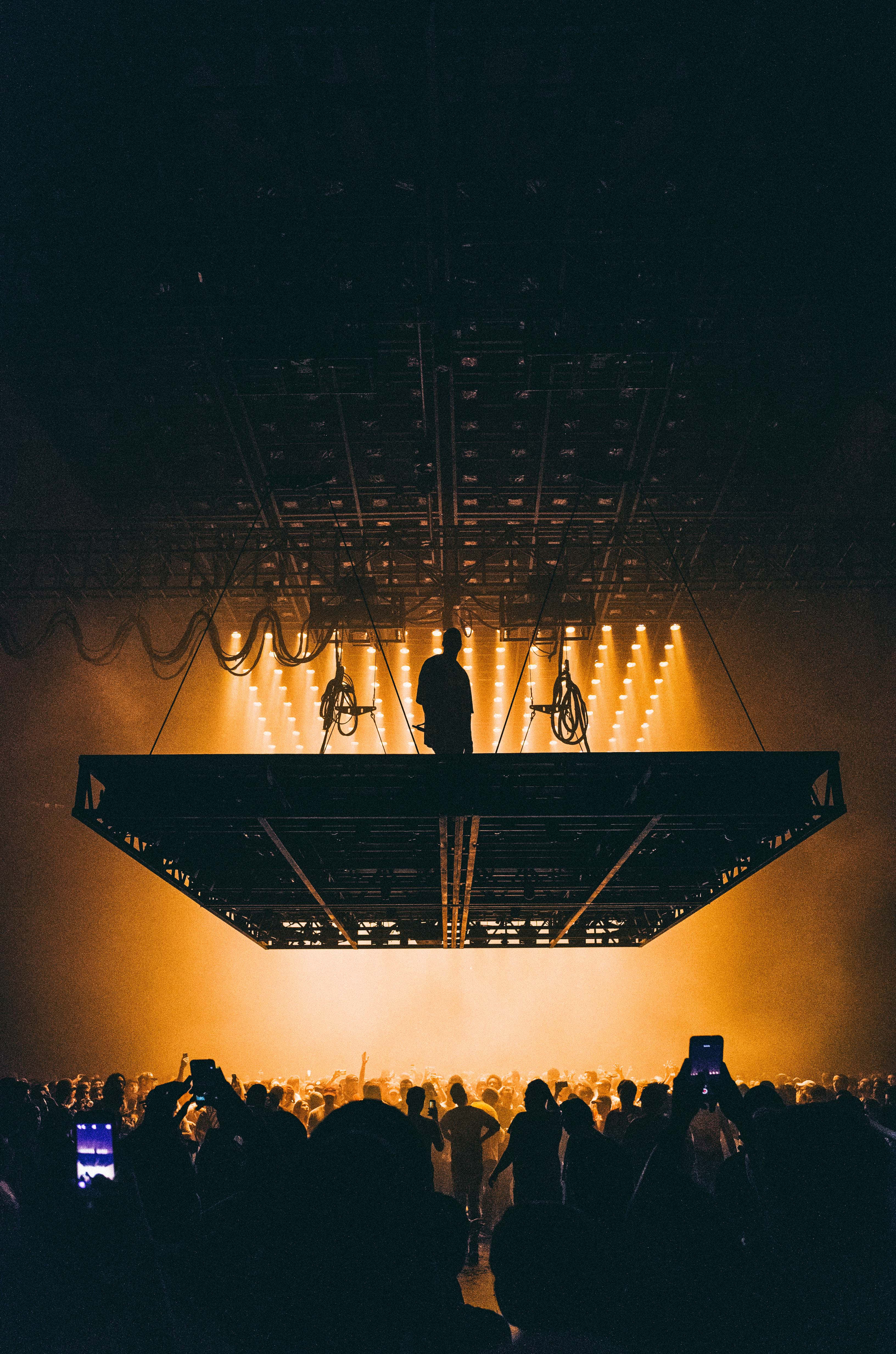 坎耶·威斯特《The Life Of Pablo》專輯封面 壁紙 — 一位孤獨的表演者站在懸掛的舞台上，剪影映襯著沐浴在金色舞台燈光下的喧囂人群。