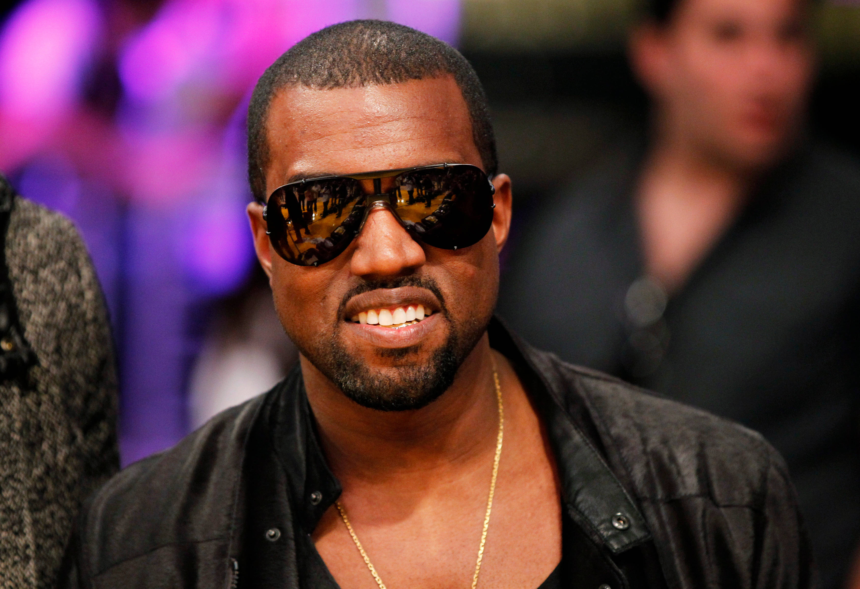 坎耶·威斯特戴飛行員太陽眼鏡 壁紙 — Kanye West 自信地微笑，戴著深色太陽眼鏡和黑色外套，背景是模糊的紫色。