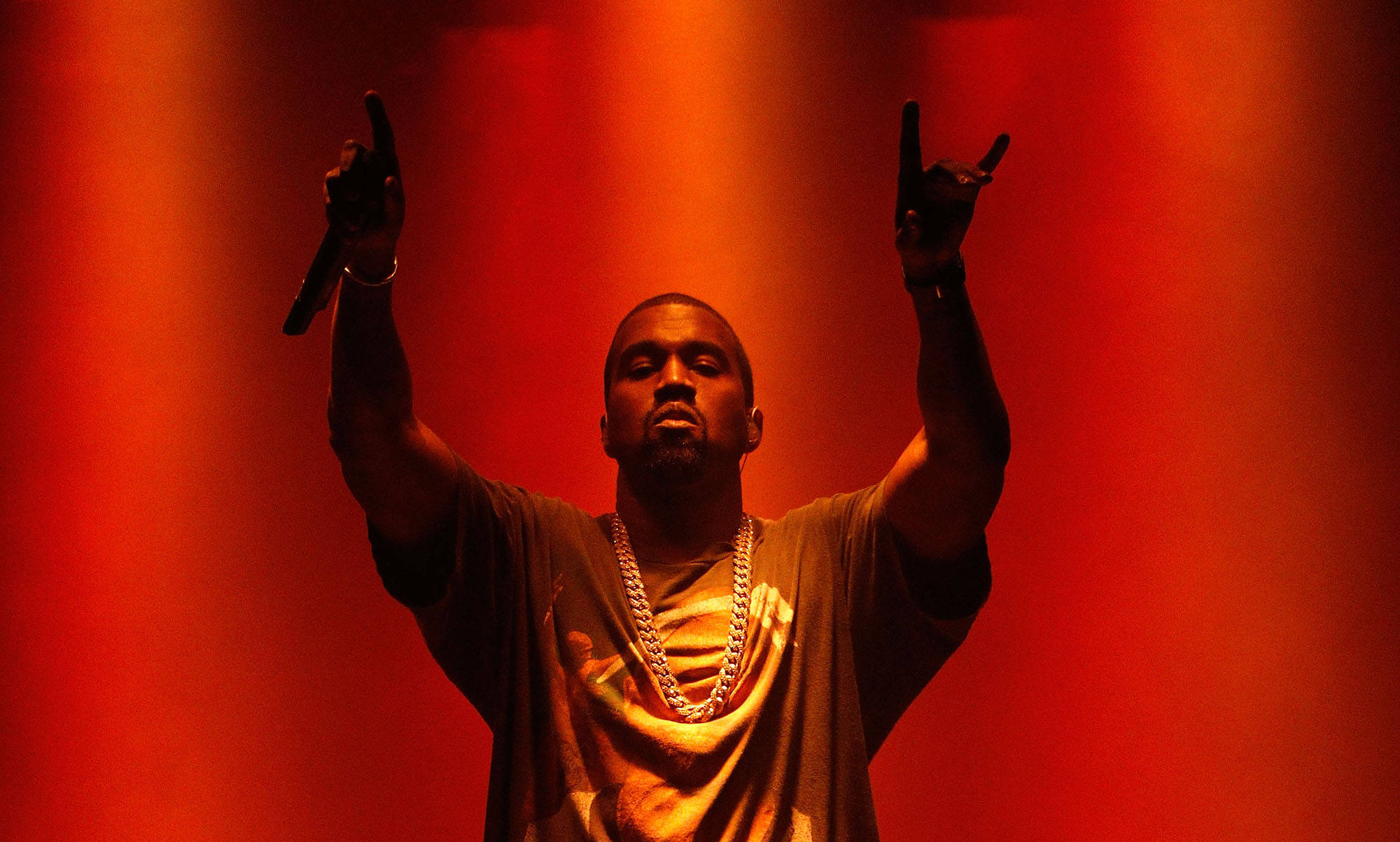 坎耶·威斯特搖滾手勢 壁紙 — Kanye West 在強烈紅燈下舉起雙臂，手持麥克風在舞台上表演。