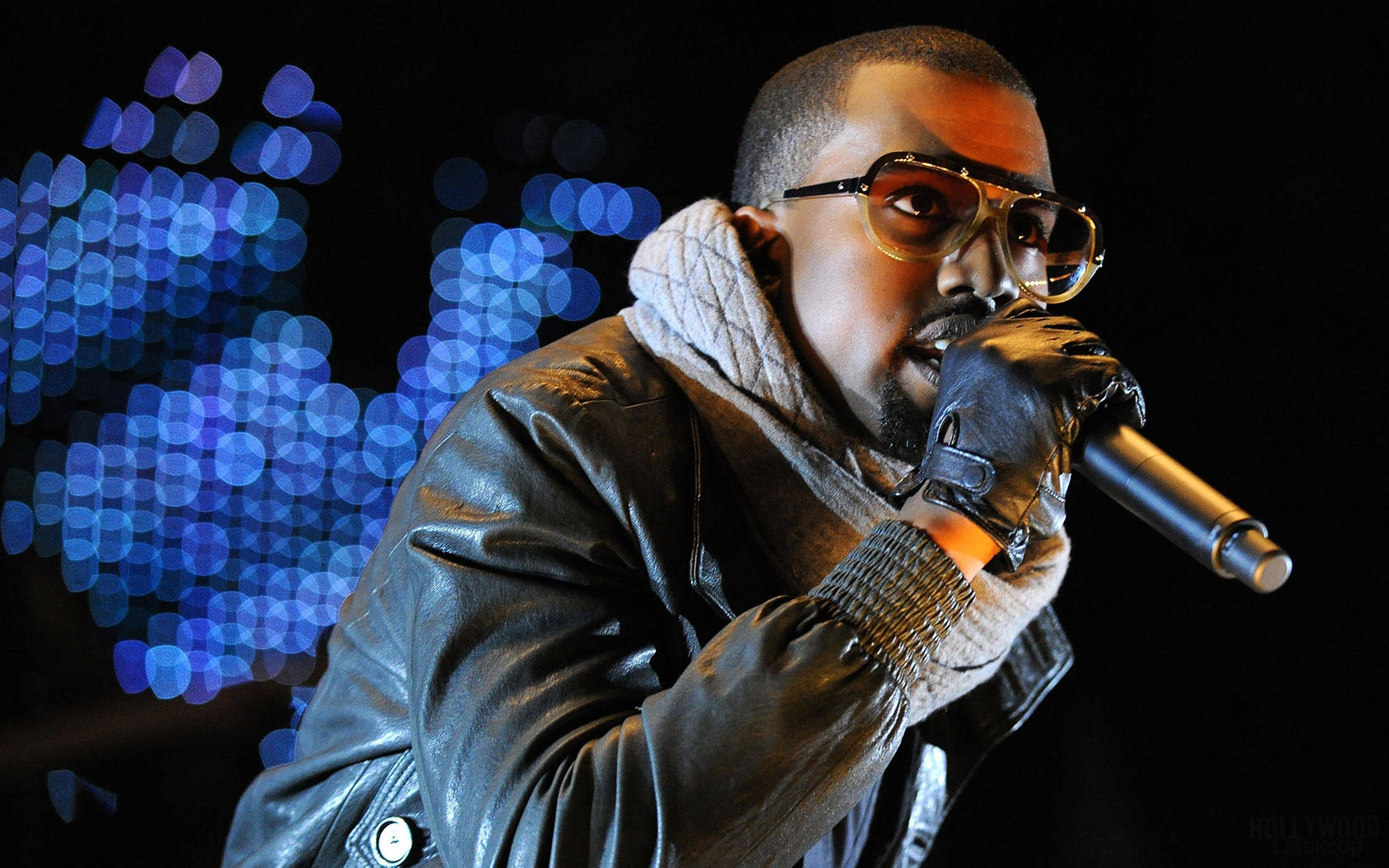 坎耶·威斯特演出現場 壁紙 — Kanye West 在舞台上表演，戴著眼鏡、皮褸和手套，手持咪高峰，背景是藍色散景燈光。