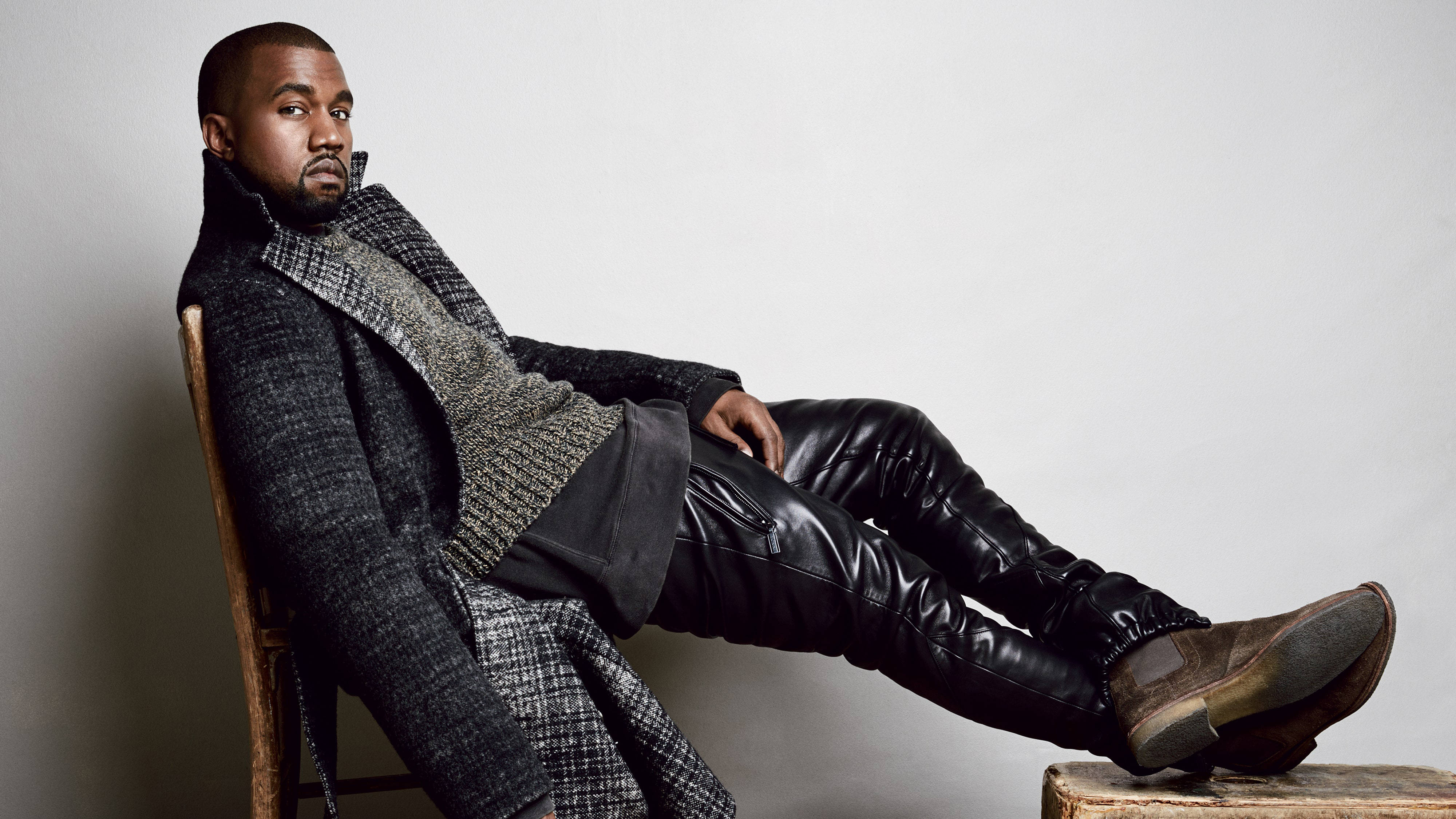 坎耶·威斯特穿黑色皮褲 壁紙 — Kanye West 自信地坐在椅子上，穿著質感外套和皮褲，背景是純灰色牆壁。