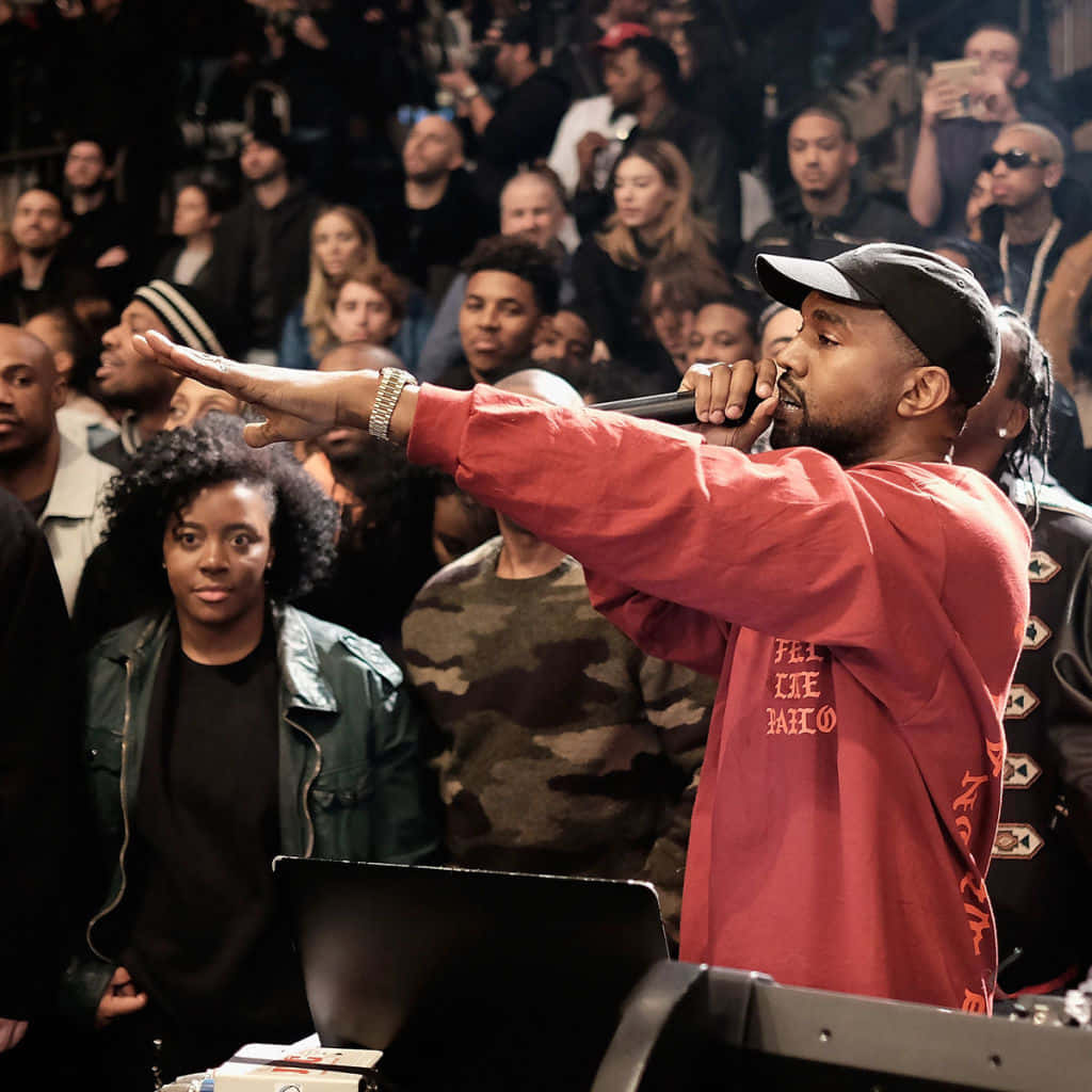 坎耶·威斯特第七張錄音室專輯《The Life Of Pablo》 壁紙 — Kanye West 穿著紅色襯衫和黑色帽子，在昏暗的場地中熱情地對著麥克風表演，同時指向人群。