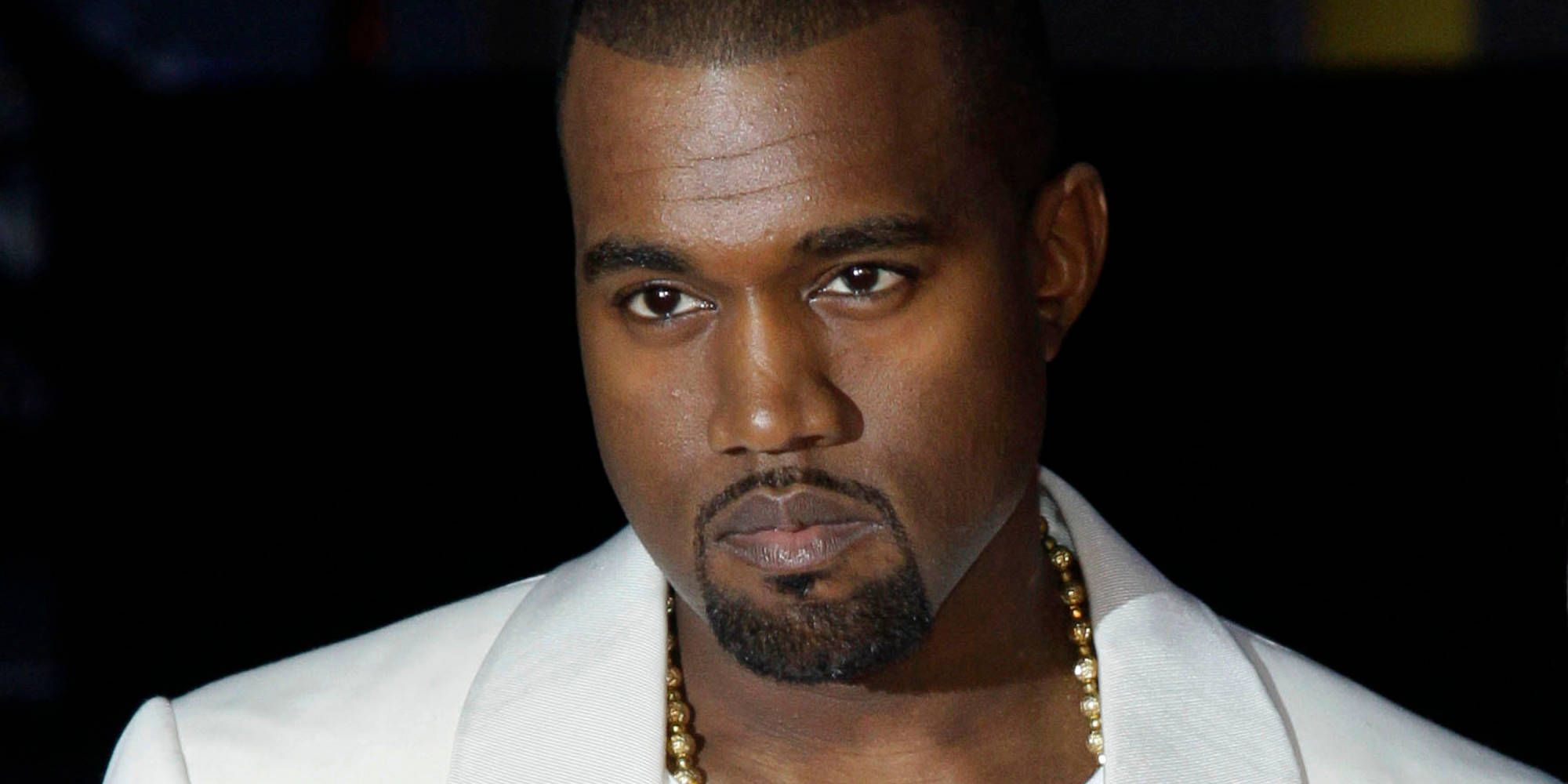 坎耶·威斯特美國饒舌歌手 壁紙 — Kanye West 身穿白色西裝和金色鏈條，在深色背景下的特寫肖像。