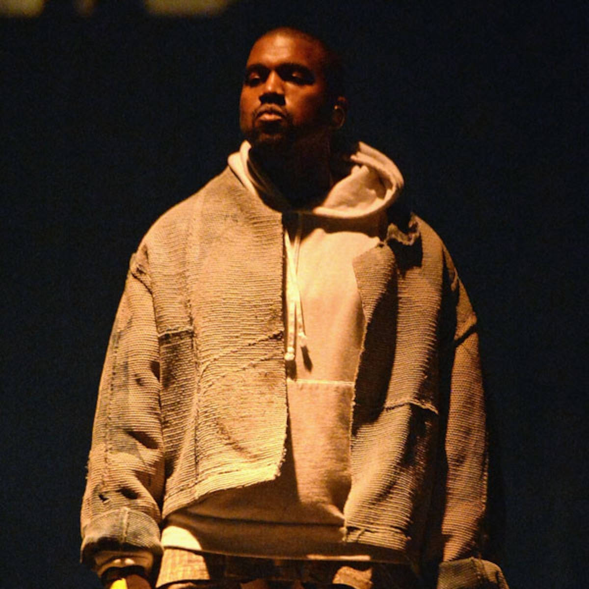 Kanye West 穿著質感米色連帽衫，在聚光燈下的黑暗舞台上。