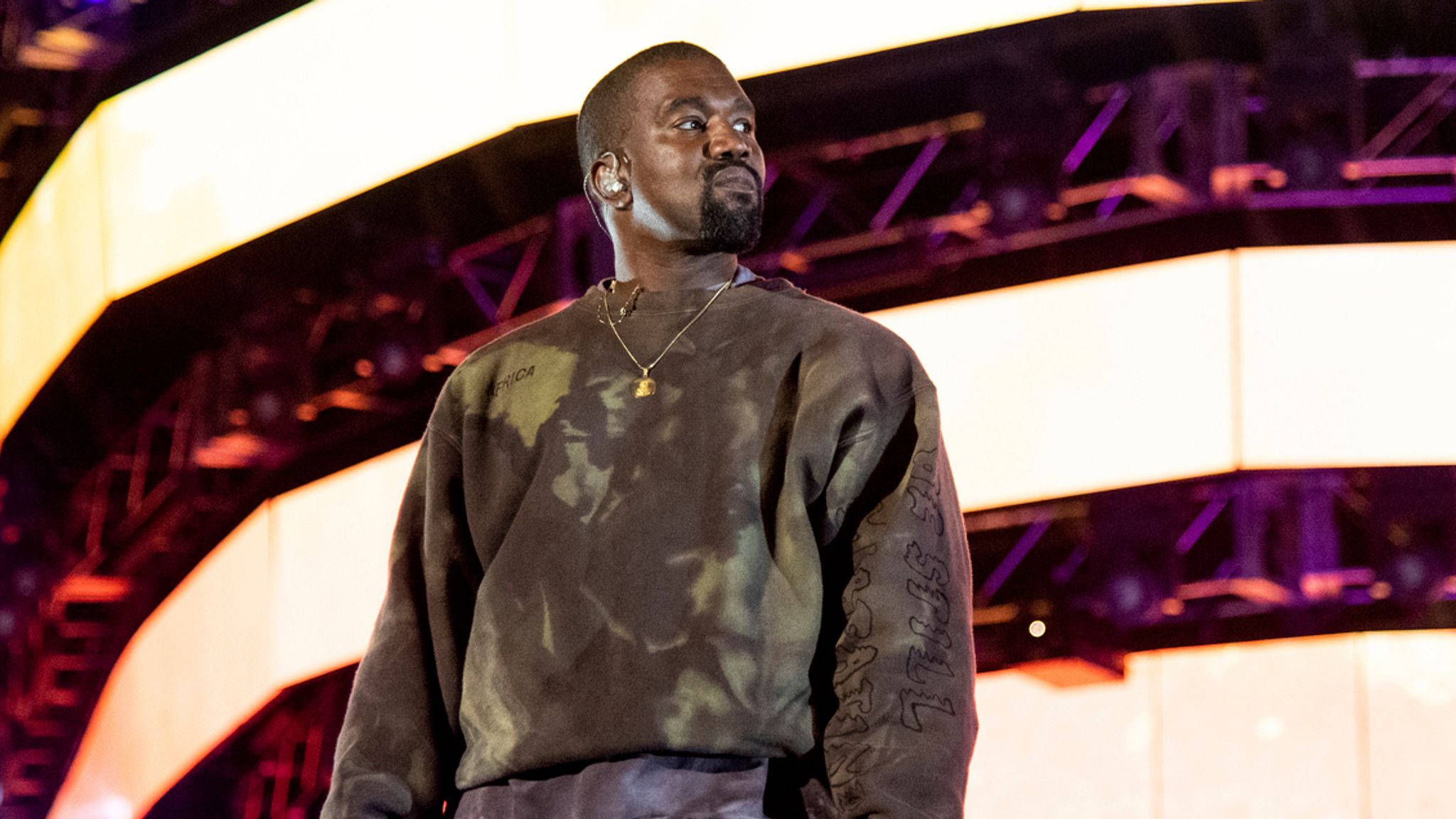坎耶·威斯特製作母親Donda West的遺作專輯 壁紙 — Kanye West 站在燈光明亮的舞台上，在彩色舞台燈光下穿著深色紮染運動衫。