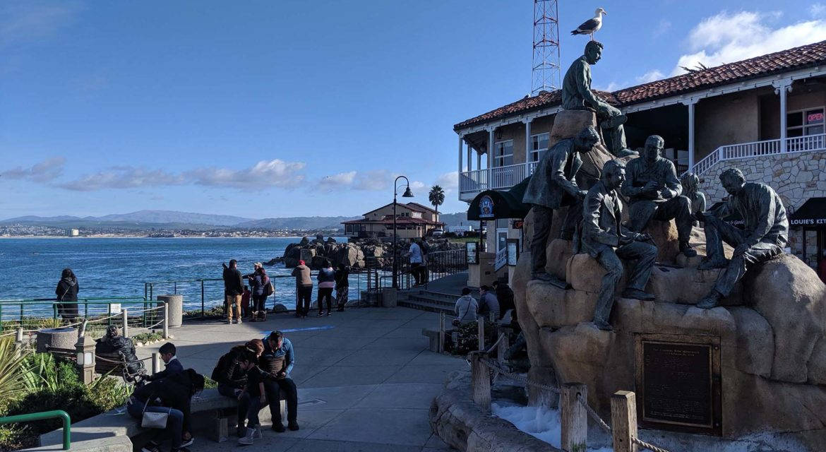 坎農行街紀念雕像 壁紙 — 一座漁夫銅像棲息在海邊步道旁的石頭上，人們在晴朗的藍天下放鬆。