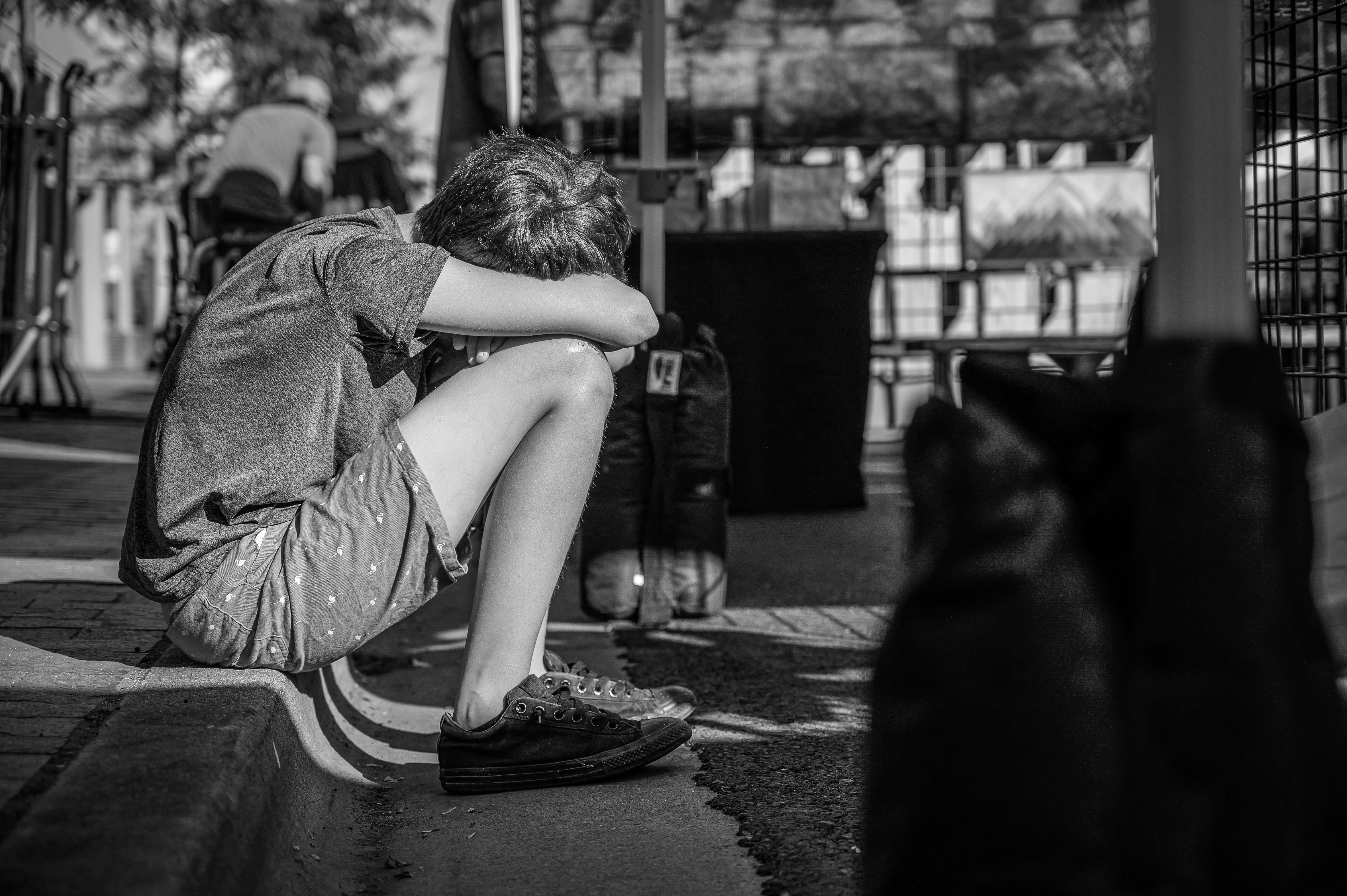坐在人行道上的憂鬱男孩 壁紙 — 一張黑白照片，拍攝一個孩子在城市環境中蜷縮在路邊，頭埋在膝蓋裡。