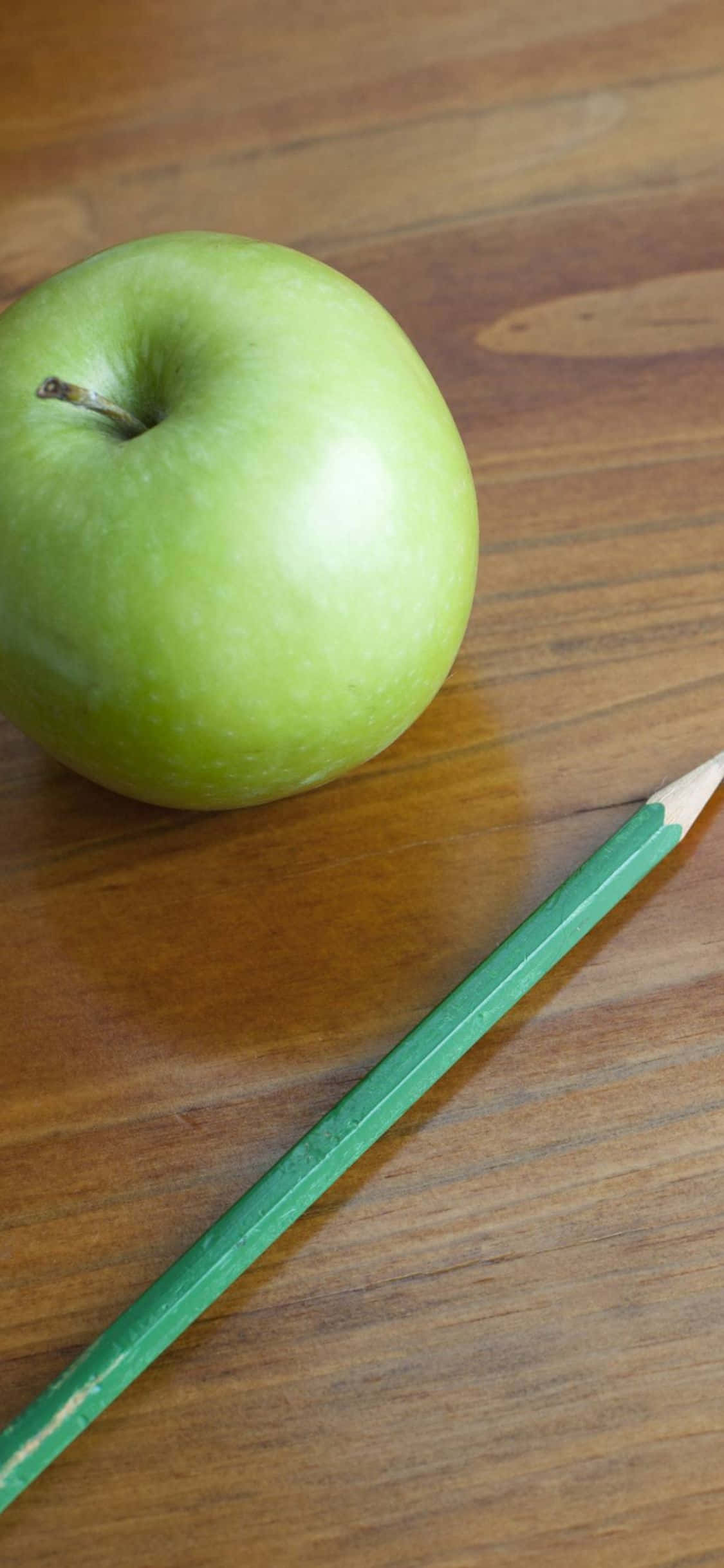 坐在書桌前，使用我的新款 iPhone XS 壁紙 — 一個清脆的青蘋果放在溫暖的木質表面上，旁邊有一支綠色鉛筆。