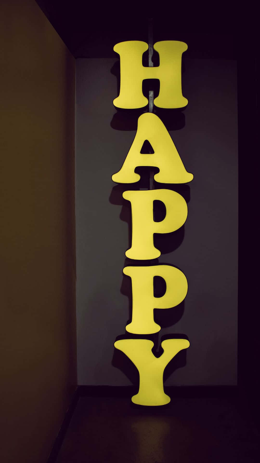 Happy iPhone 桌布 壁紙 — 垂直的黃色「HAPPY」字母在昏暗的室內角落中，在深色牆壁上溫暖地發光。