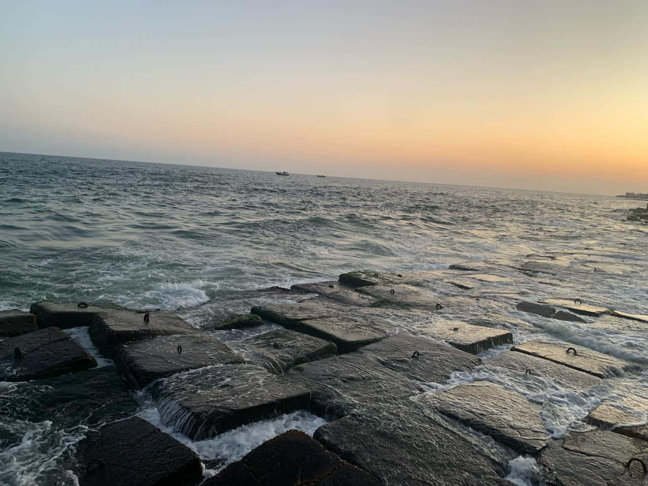 海浪拍打石堤，夕陽下遠方船隻在平靜海平線上