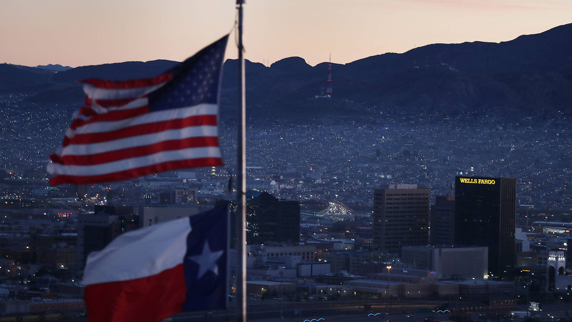 美國國旗和德克薩斯州旗在黃昏時分於城市上空飄揚，背後有山脈。