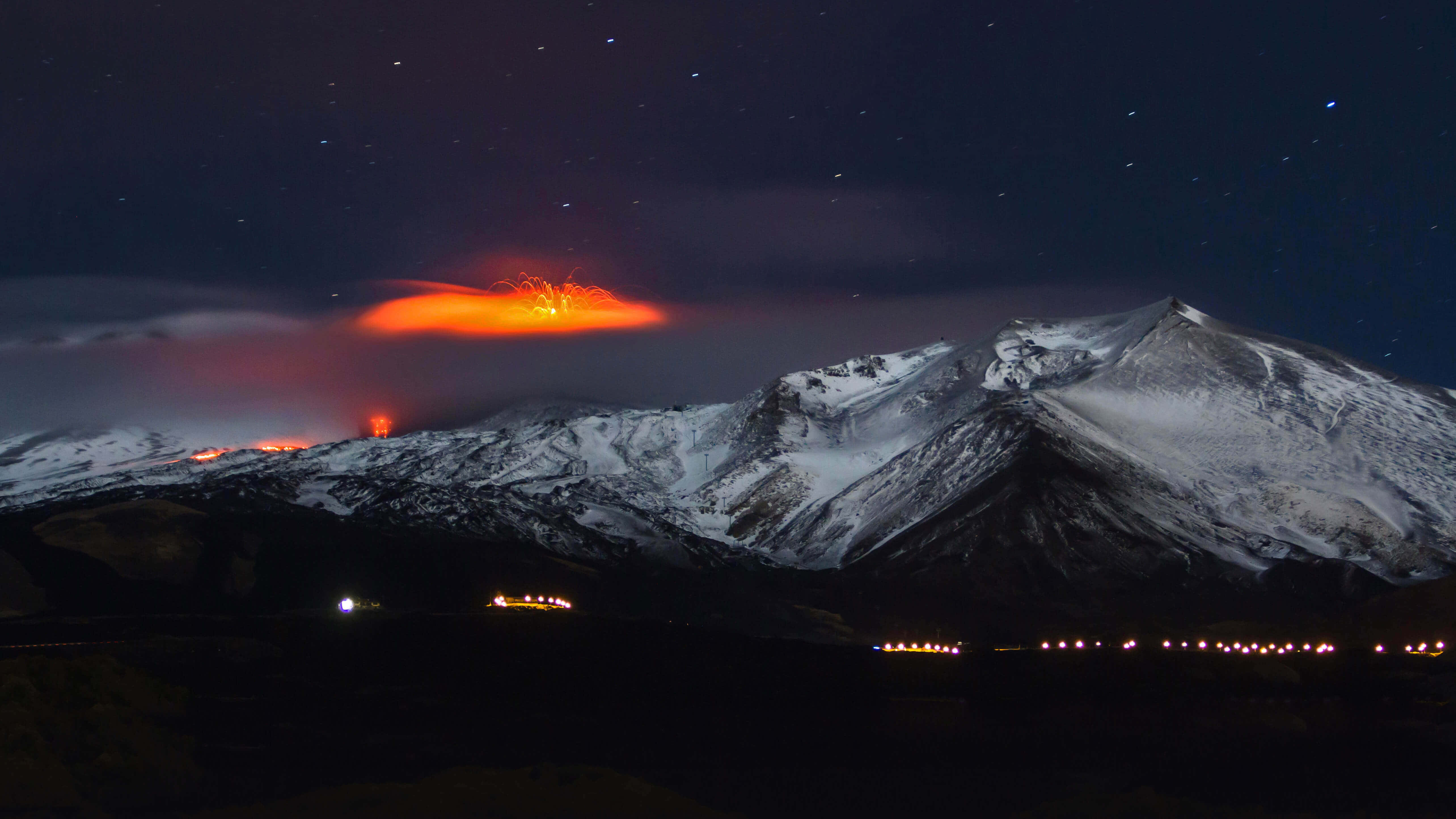 埃特納火山夜景暗光海報 壁紙 — 被雪覆蓋的山脈在繁星點點的夜空下噴出熾熱的橙色熔岩，下方有遠處的燈光。