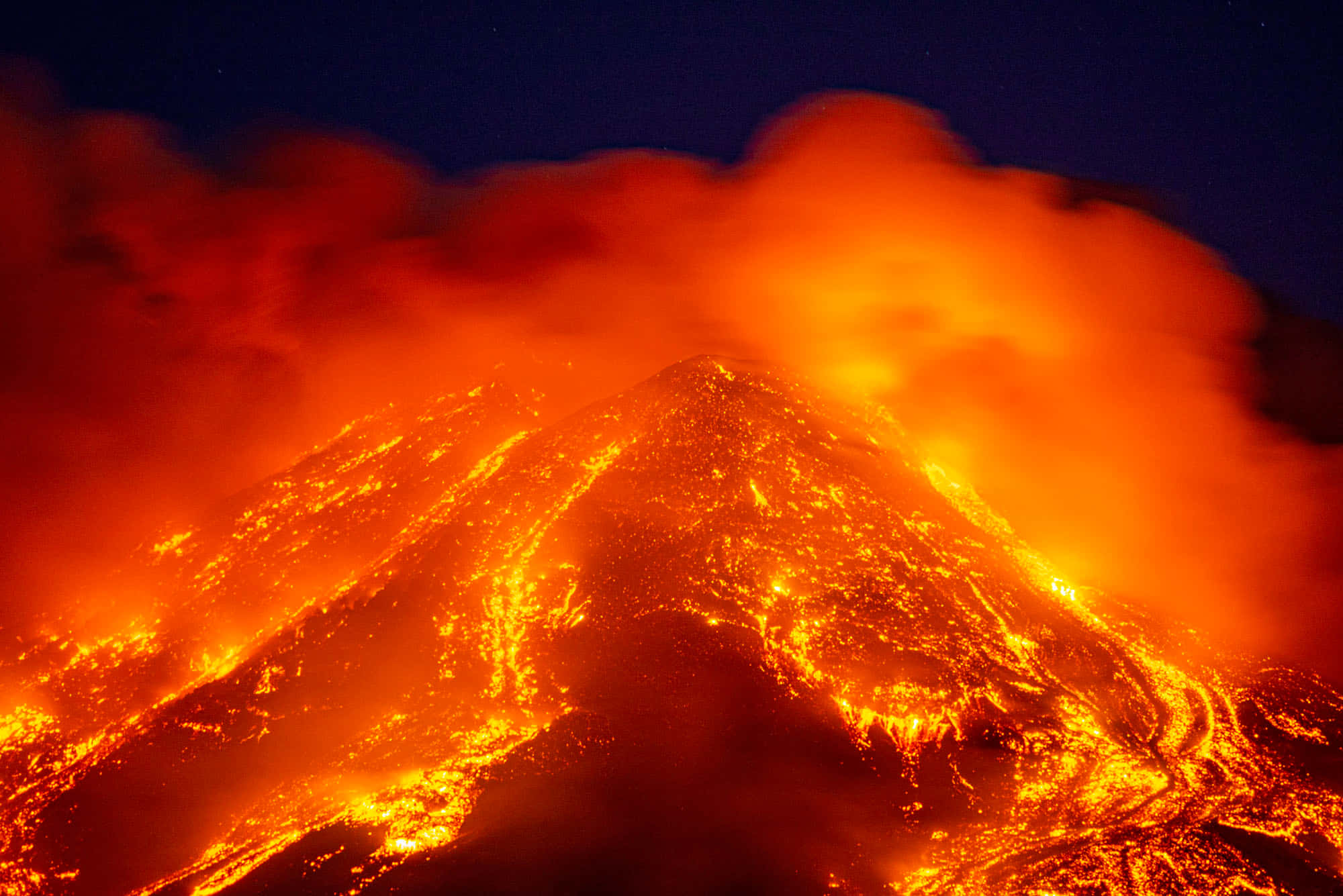 埃特納火山氣候火山 壁紙 — 一座火山在夜間爆發，發光的橙色熔岩在黑暗的天空下沿著山坡流下。
